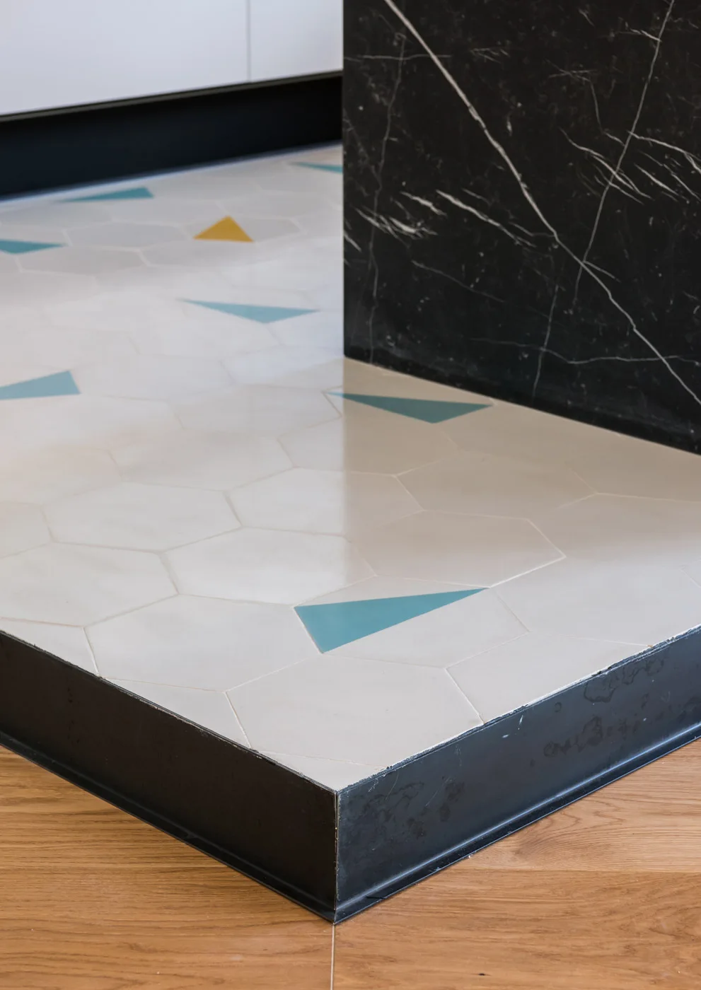 White hexagonal floor tiles with teal triangle accents transition to light wood flooring with a black metal trim strip next to a black marble island.