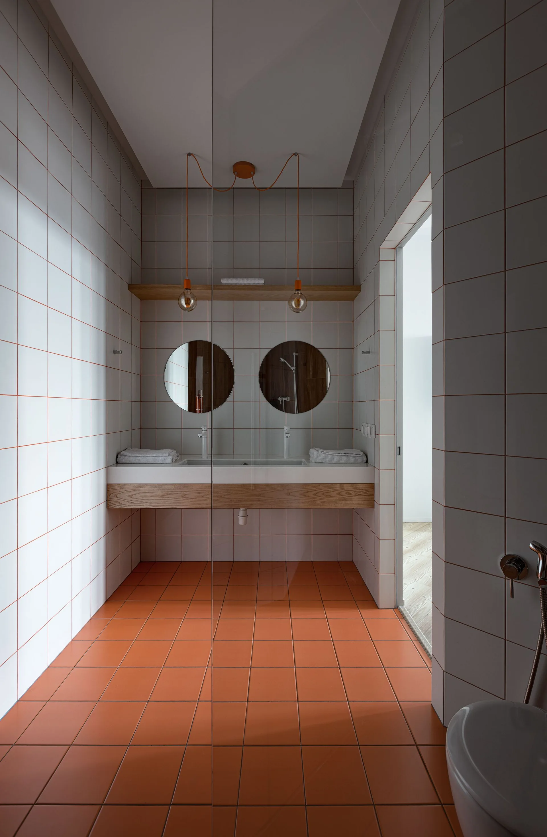 A minimalist bathroom features bright orange floor tiles, white square wall tiles with matching orange grout, and a floating wood vanity with circular mirrors.
