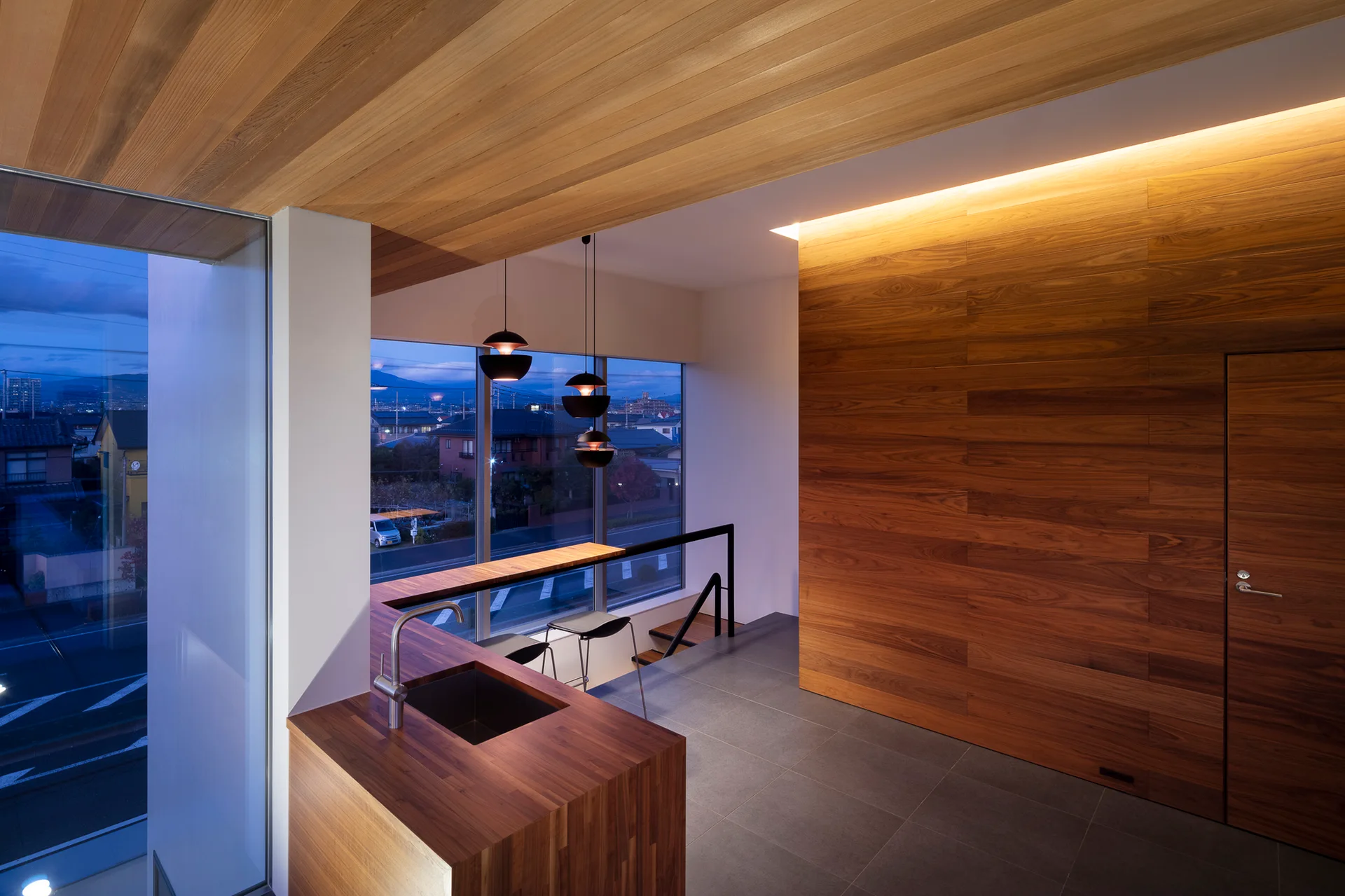 A modern kitchen features warm wood paneling on the walls and ceiling, a gray tiled floor, a matching wood island with a sink, and black pendant lights.