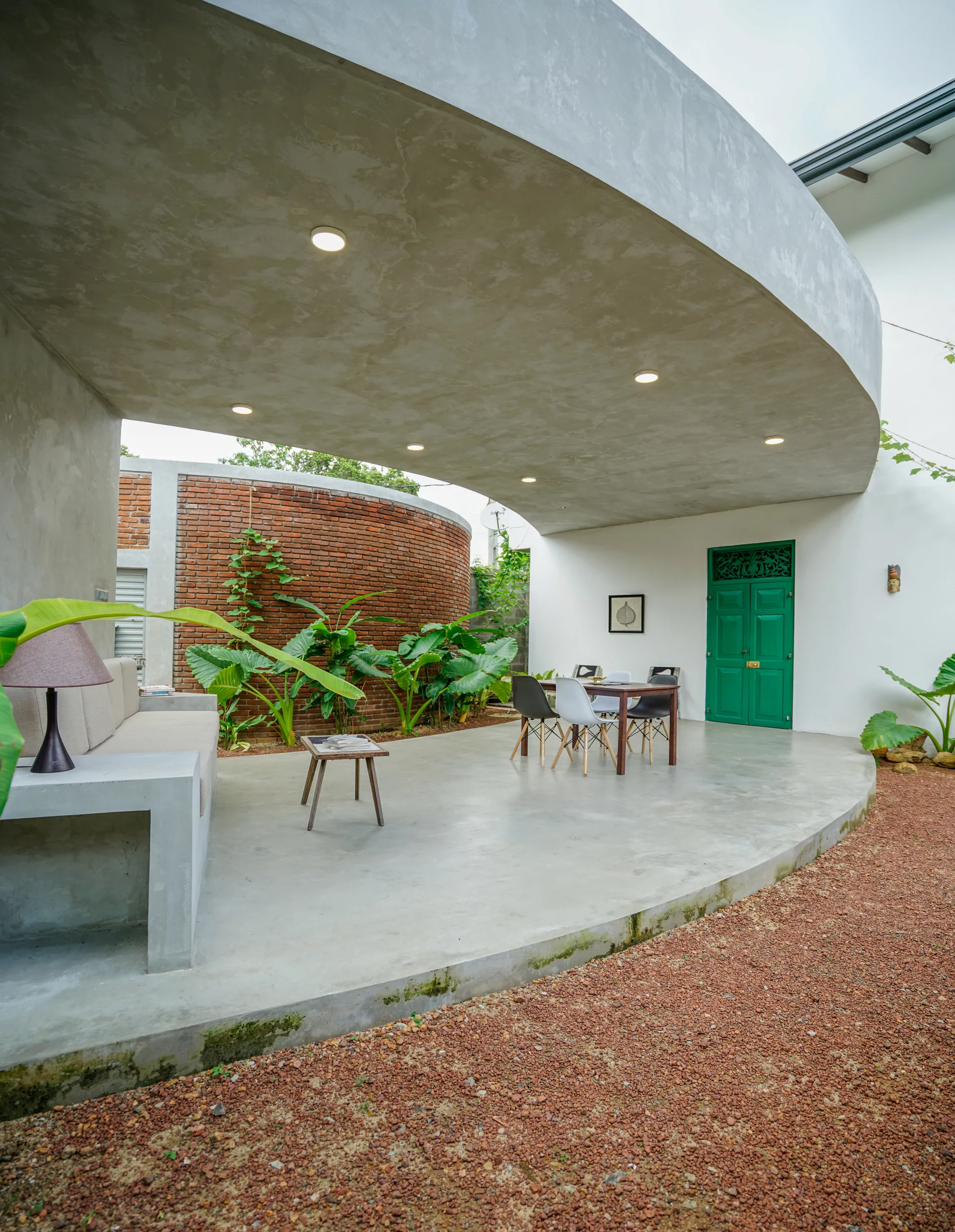An outdoor covered concrete patio features a curved overhang, a curved red brick wall with leafy plants, a green double door, and a dining set.