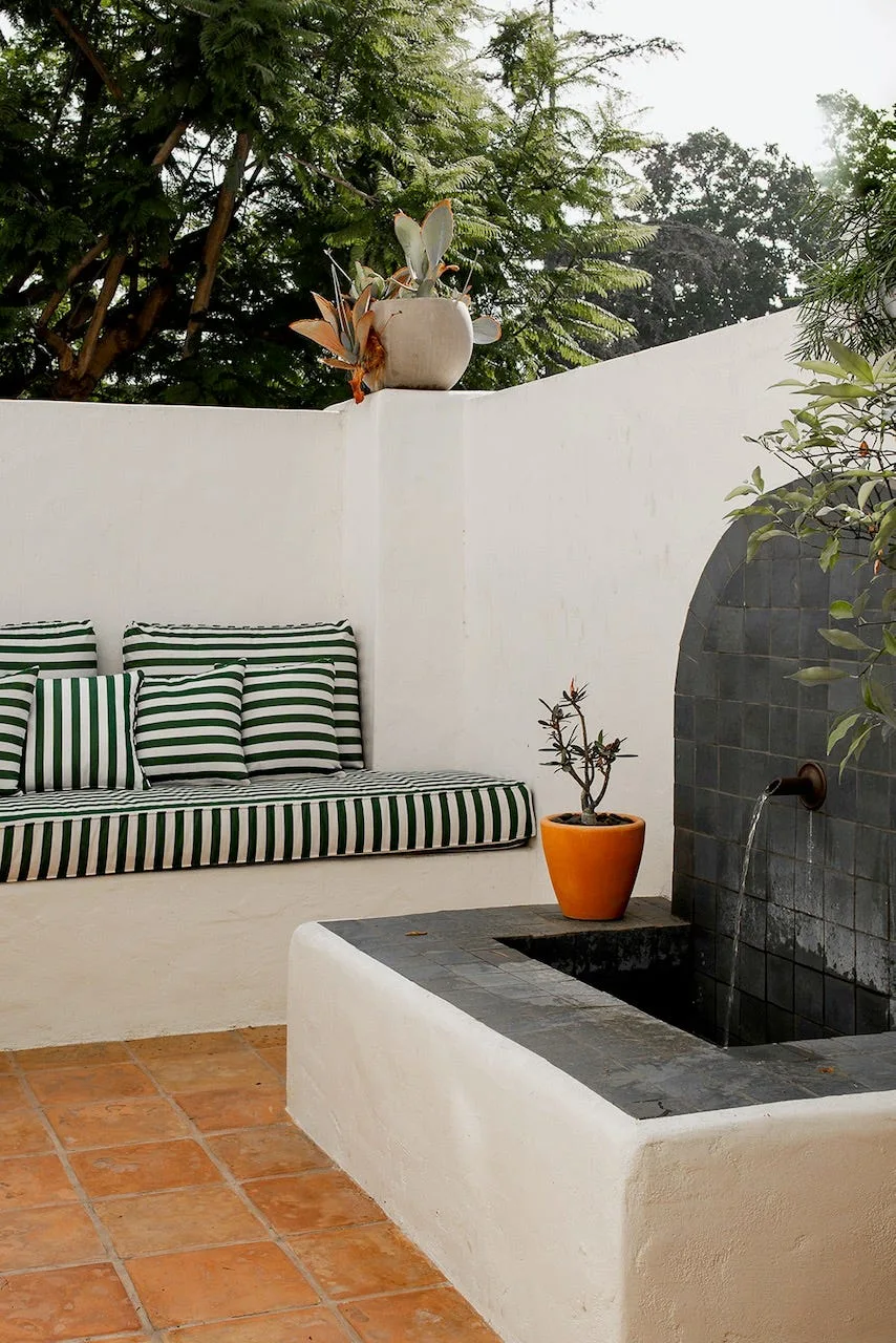 A white stucco built-in patio bench with green striped cushions sits next to a dark-tiled water fountain on a terracotta tiled floor.