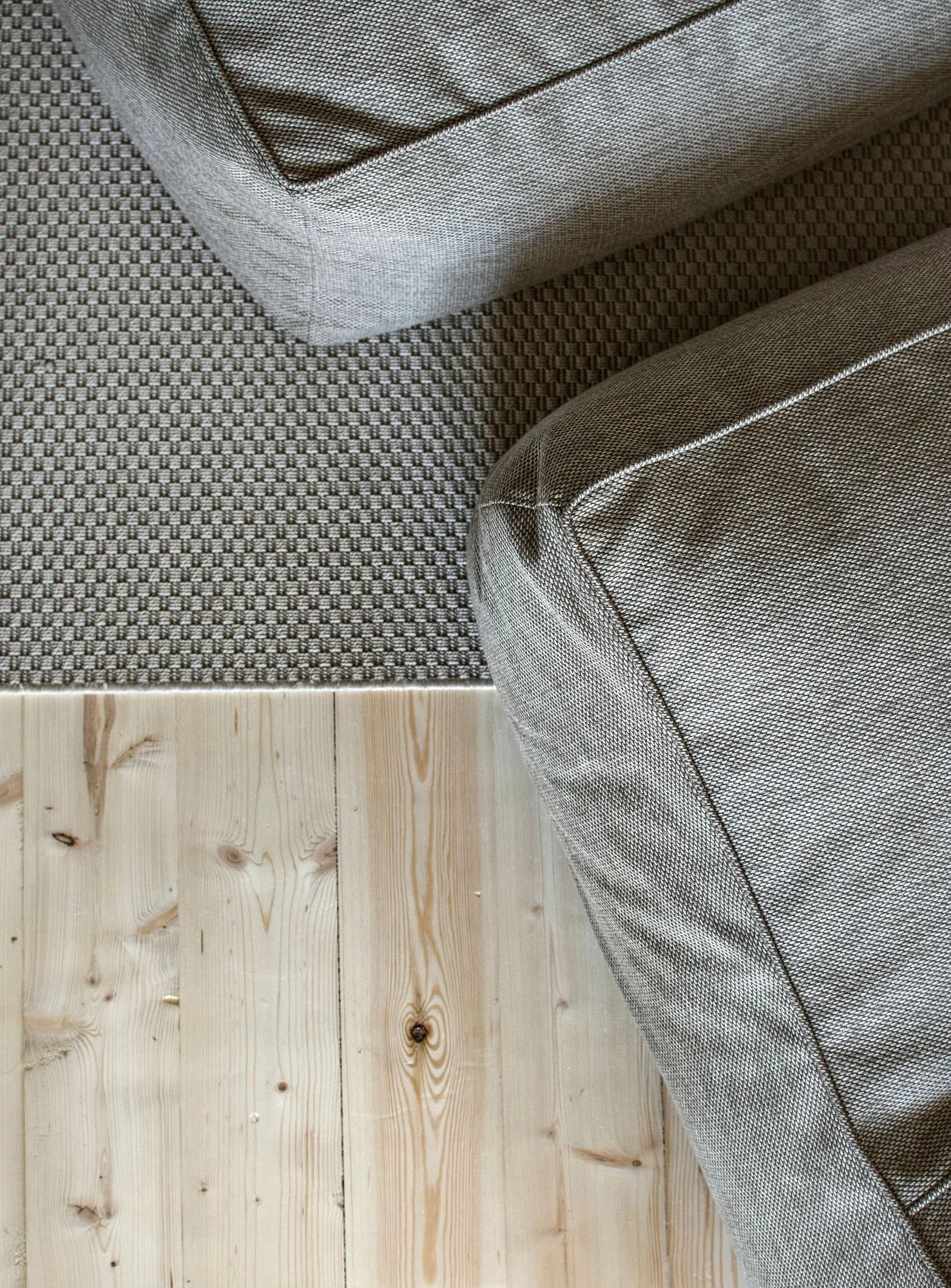A top-down close-up view of a textured gray upholstered sofa corner resting on a woven beige area rug beside light pine wood floorboards.
