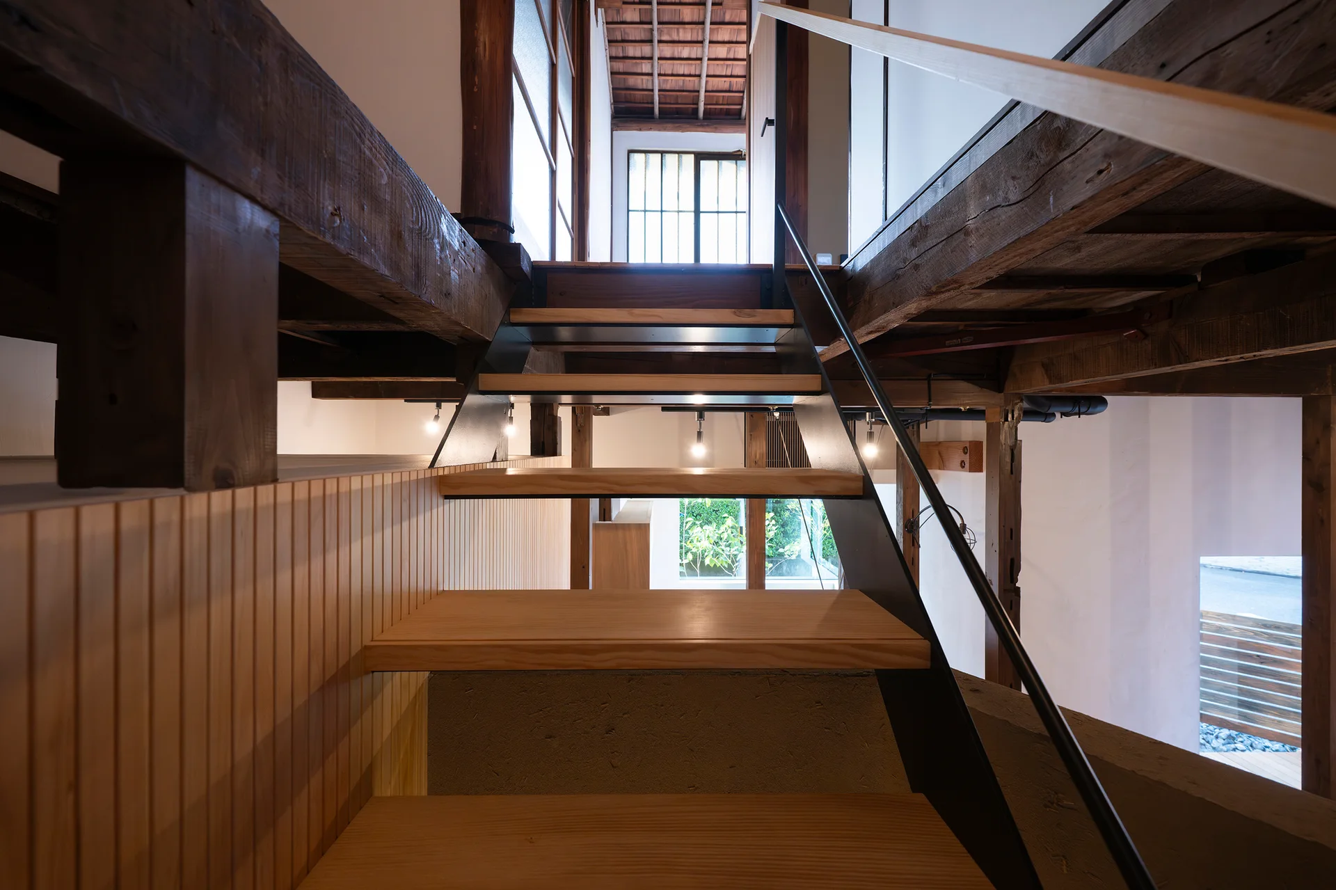 An interior staircase with light wood treads and a black metal railing rises beneath historic dark timber beams and alongside a slatted wood wall.