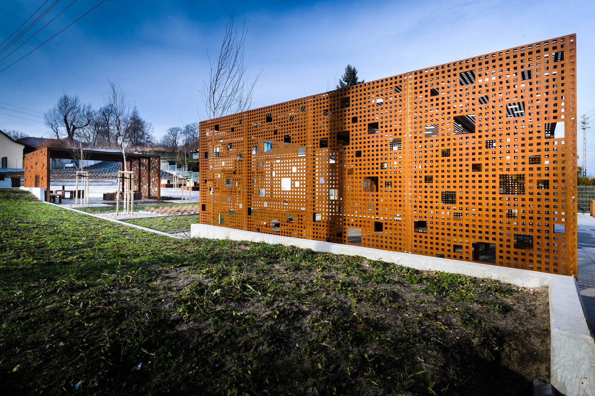A weathered rust-colored perforated metal privacy screen sits on a concrete base next to a grassy lawn with a wooden pergola in the background.