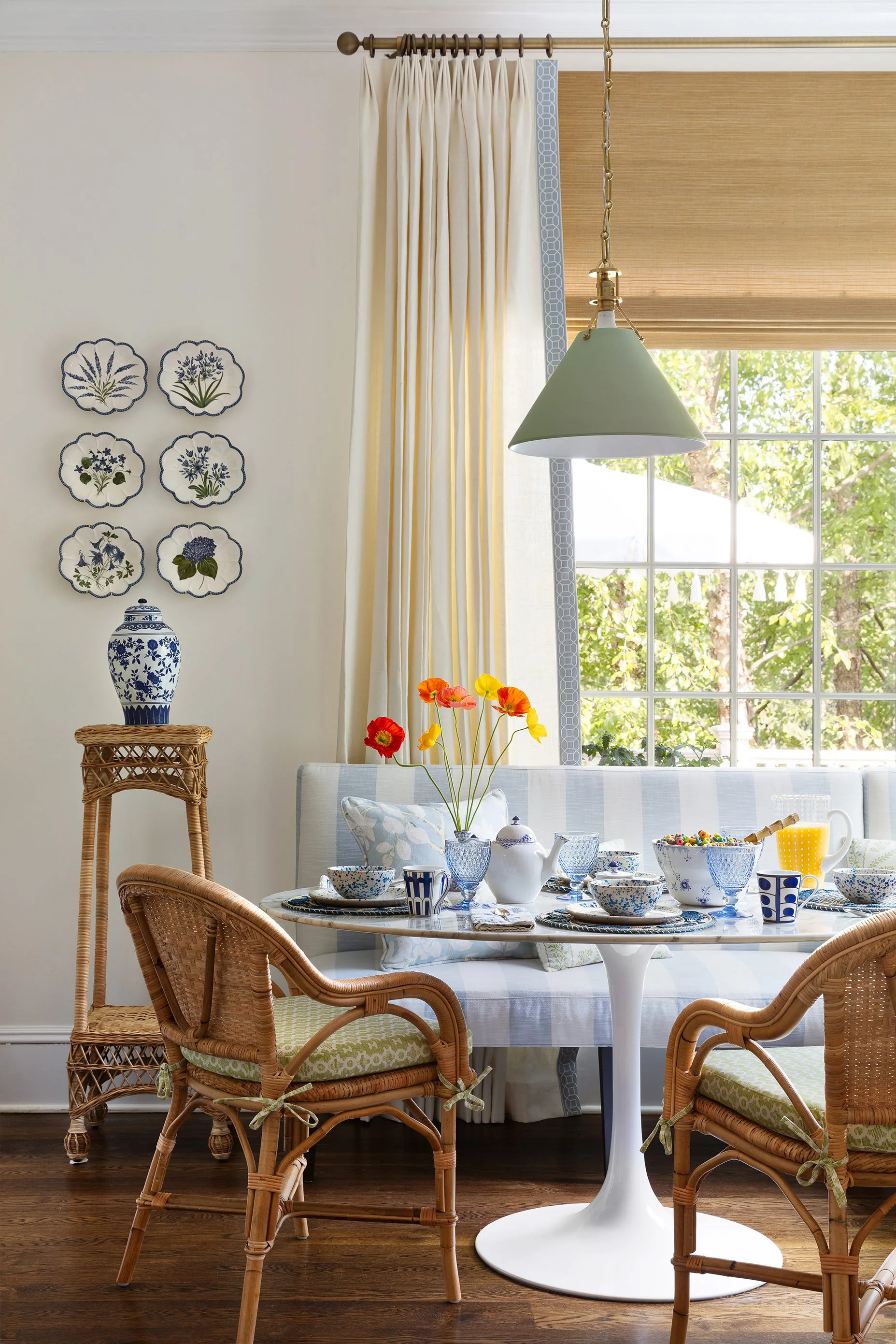 A breakfast nook features a circular marble tulip table, a blue and white striped banquette, and natural rattan chairs set against a window.