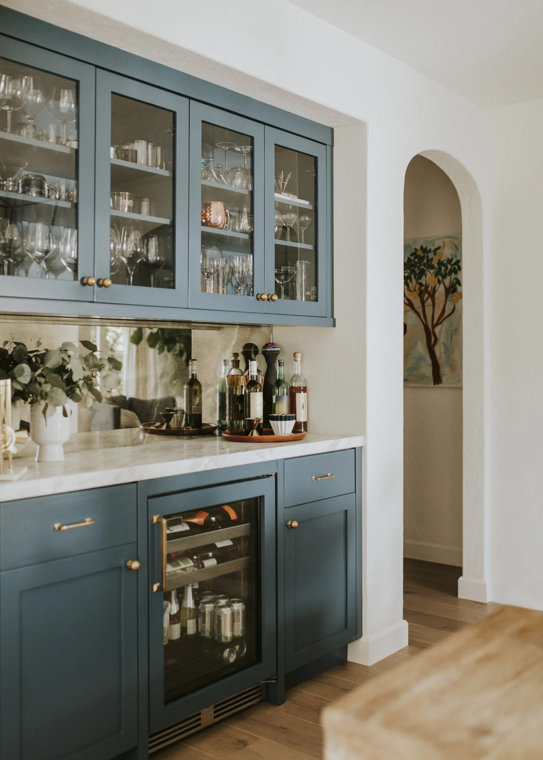 A built-in dark blue wet bar features glass-front upper cabinets, a marble countertop with bottles, a mirrored backsplash, and a wine cooler.