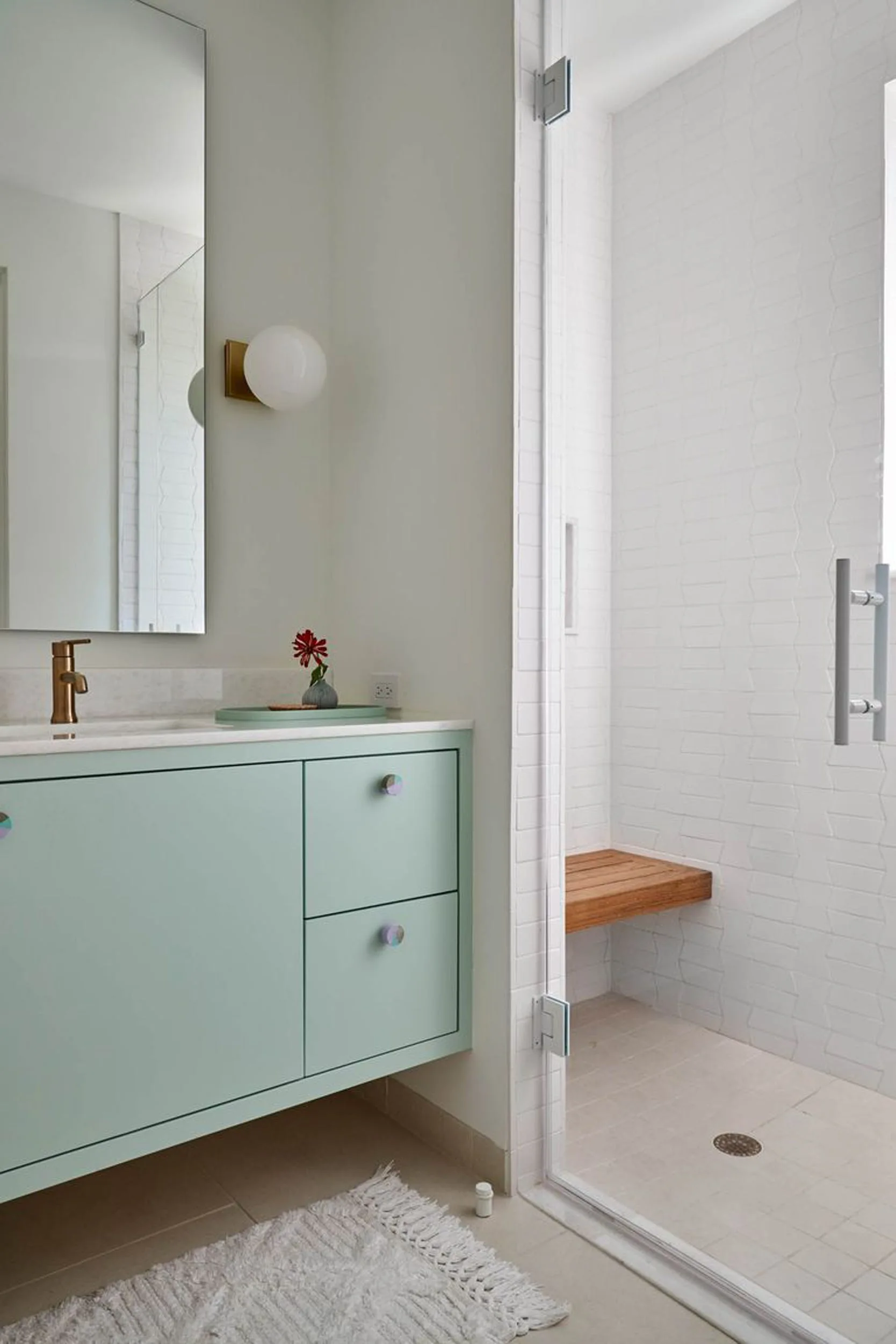A light green bathroom vanity with a brass faucet stands next to a glass-enclosed shower featuring white geometric tiles and a floating wood bench.