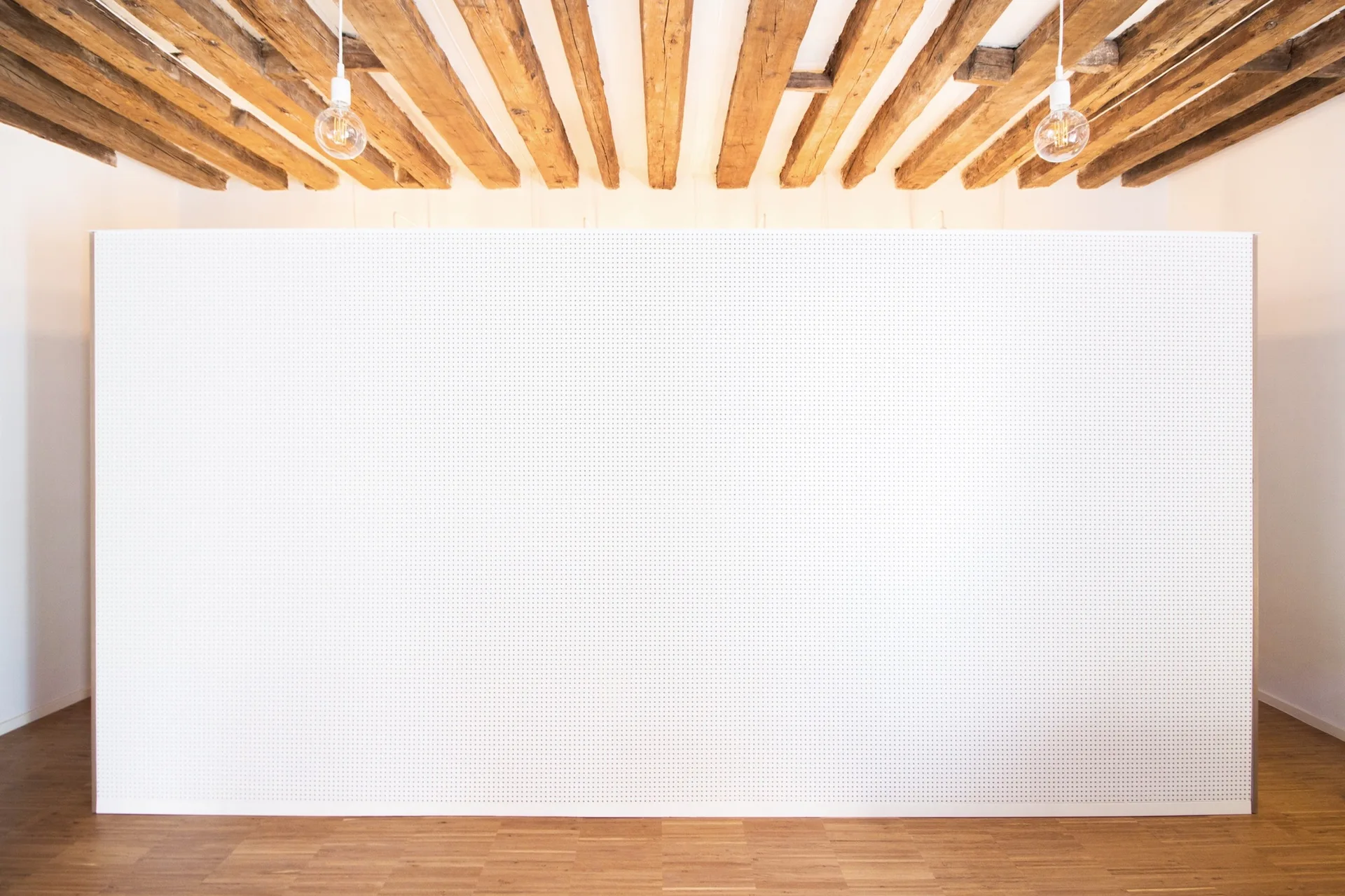 A minimalist white pegboard partition wall stands on hardwood floors beneath a ceiling of exposed natural wood beams with two bulb pendant lights.