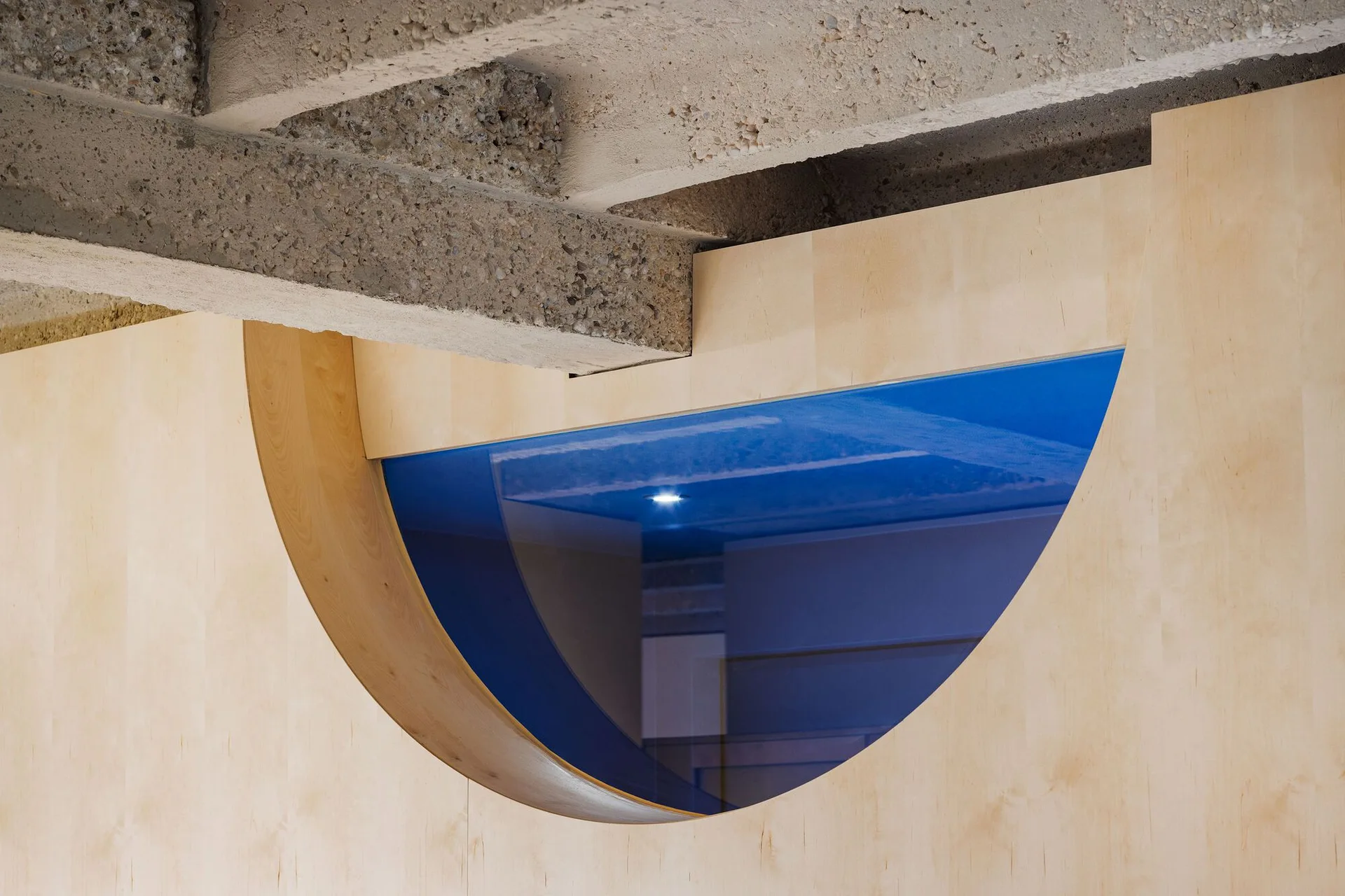 A semicircular blue glass cutout is set into a light wood wall beneath rough, textured exposed concrete ceiling beams.