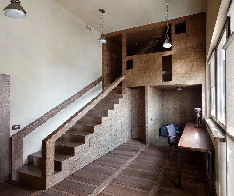 A wooden staircase with built-in storage drawers sits next to a home office nook featuring a wooden desk, chair, and large windows.