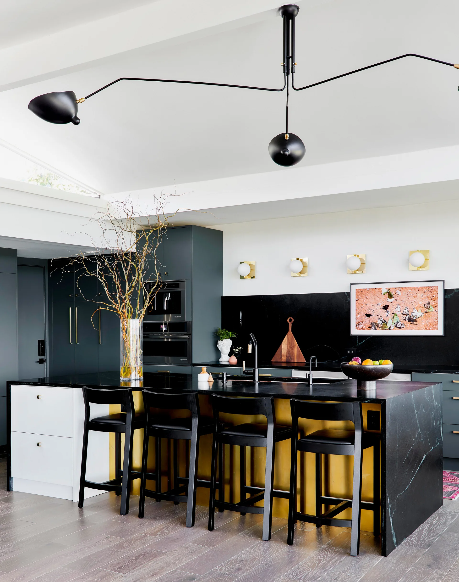A modern kitchen features a black stone waterfall island with four black stools, a gold accent base, dark green cabinets, and a black ceiling fixture.
