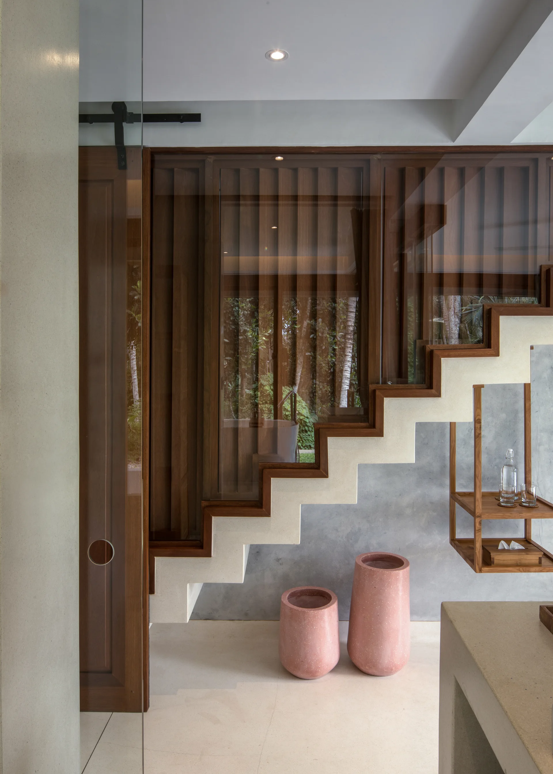 A modern staircase features zigzag beige treads trimmed in dark wood under a glass partition, with vertical wood slats and pink floor planters below.