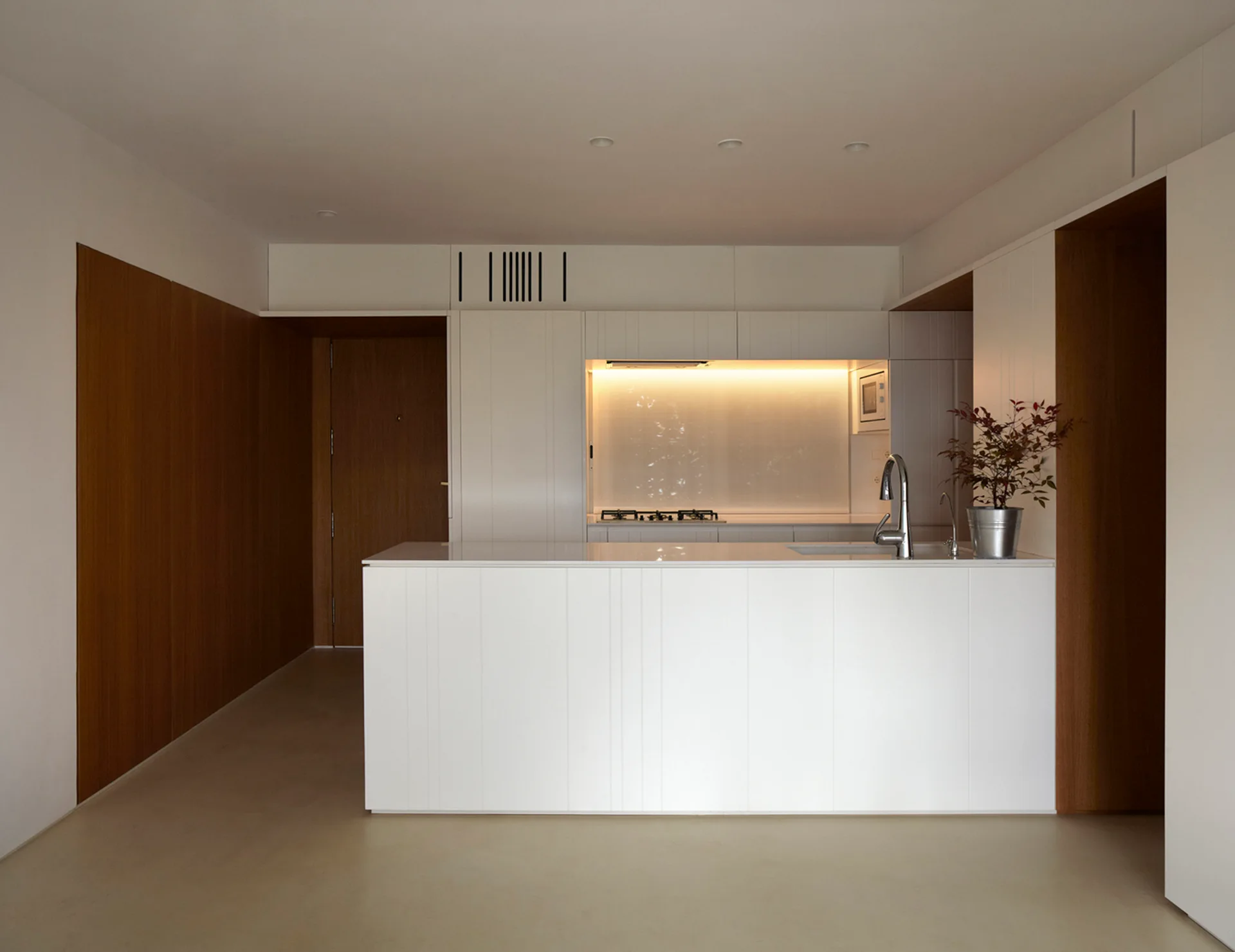 A minimalist kitchen features a white central island, flush white cabinetry with a backlit niche, and warm wood paneling on adjacent walls.