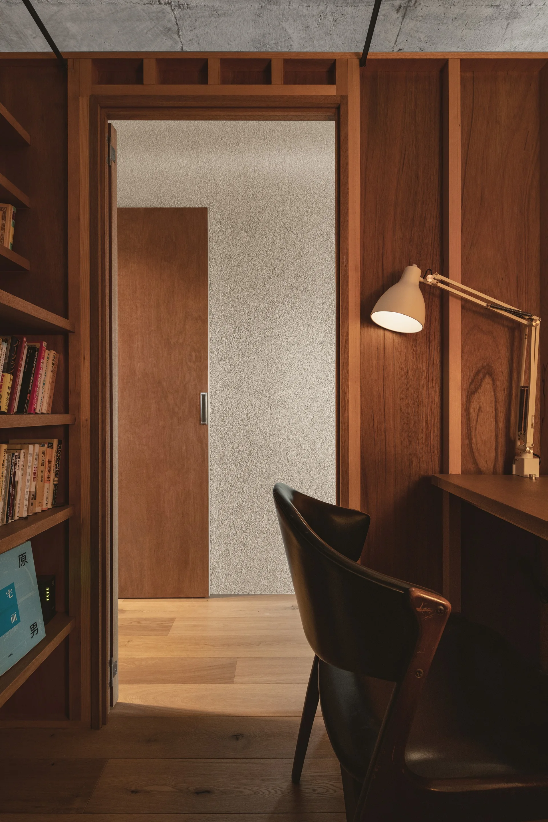 A small home office workspace with vertical wood paneling, an integrated desk with a task lamp, a leather chair, and a doorway.