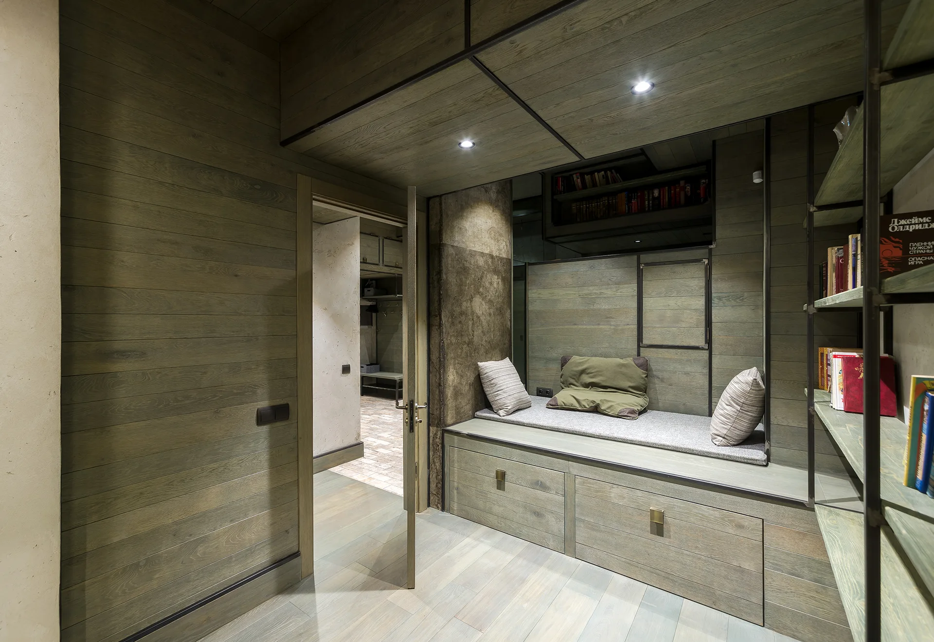 A reading nook clad in muted green wood planks features a built-in storage bench with cushions, recessed spotlights, and open metal shelving with books.