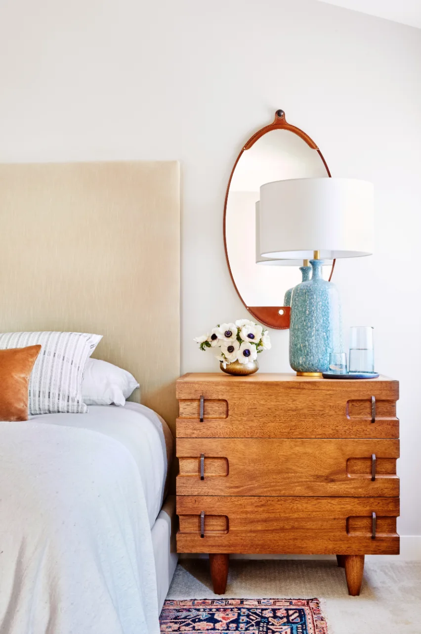 A mid-century wood nightstand with a blue speckled lamp sits next to a beige upholstered headboard below an oval wall mirror.
