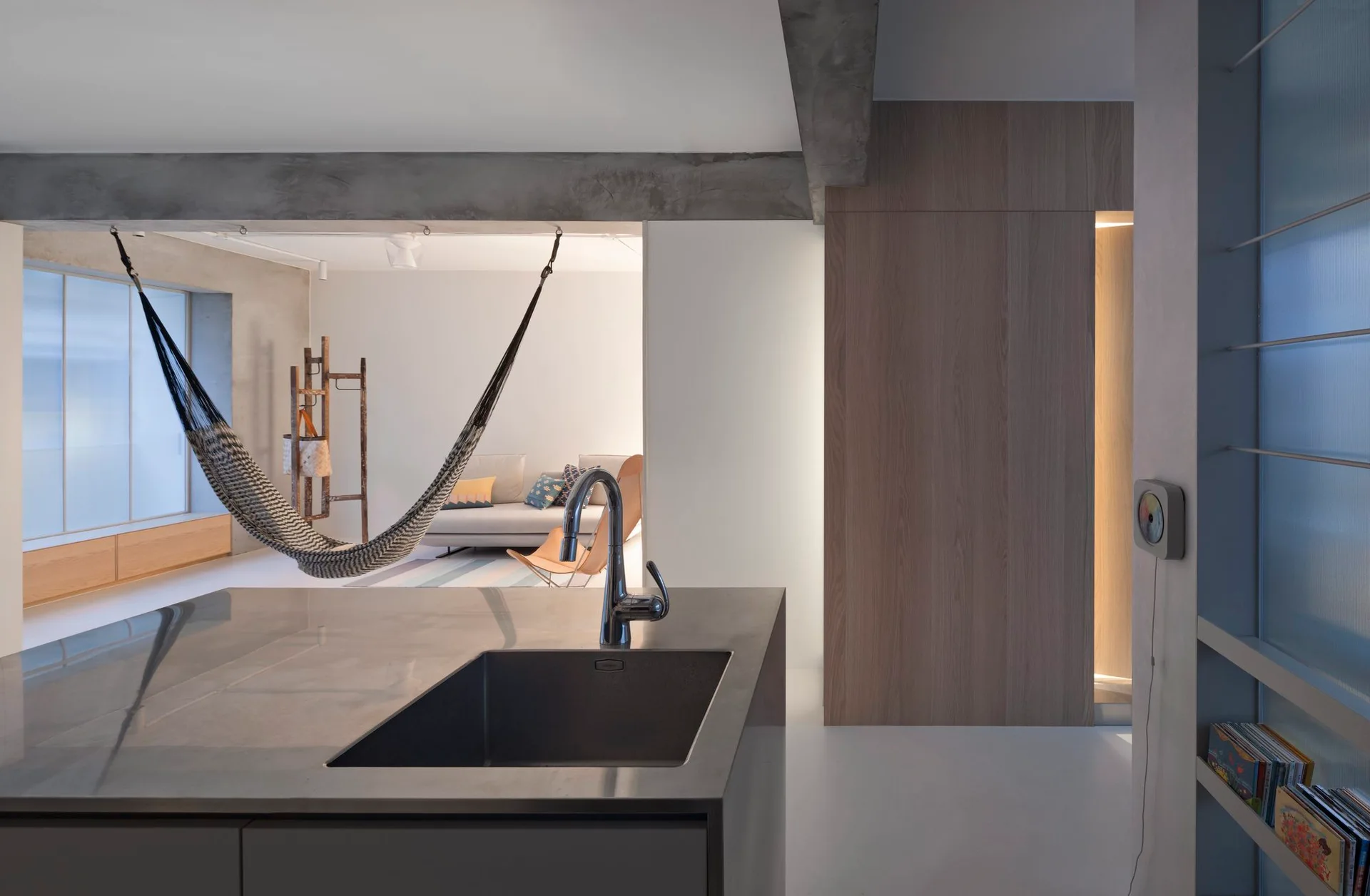 A modern grey kitchen island with a sink sits in the foreground, looking toward a white living area featuring a suspended woven hammock and a wooden wall.