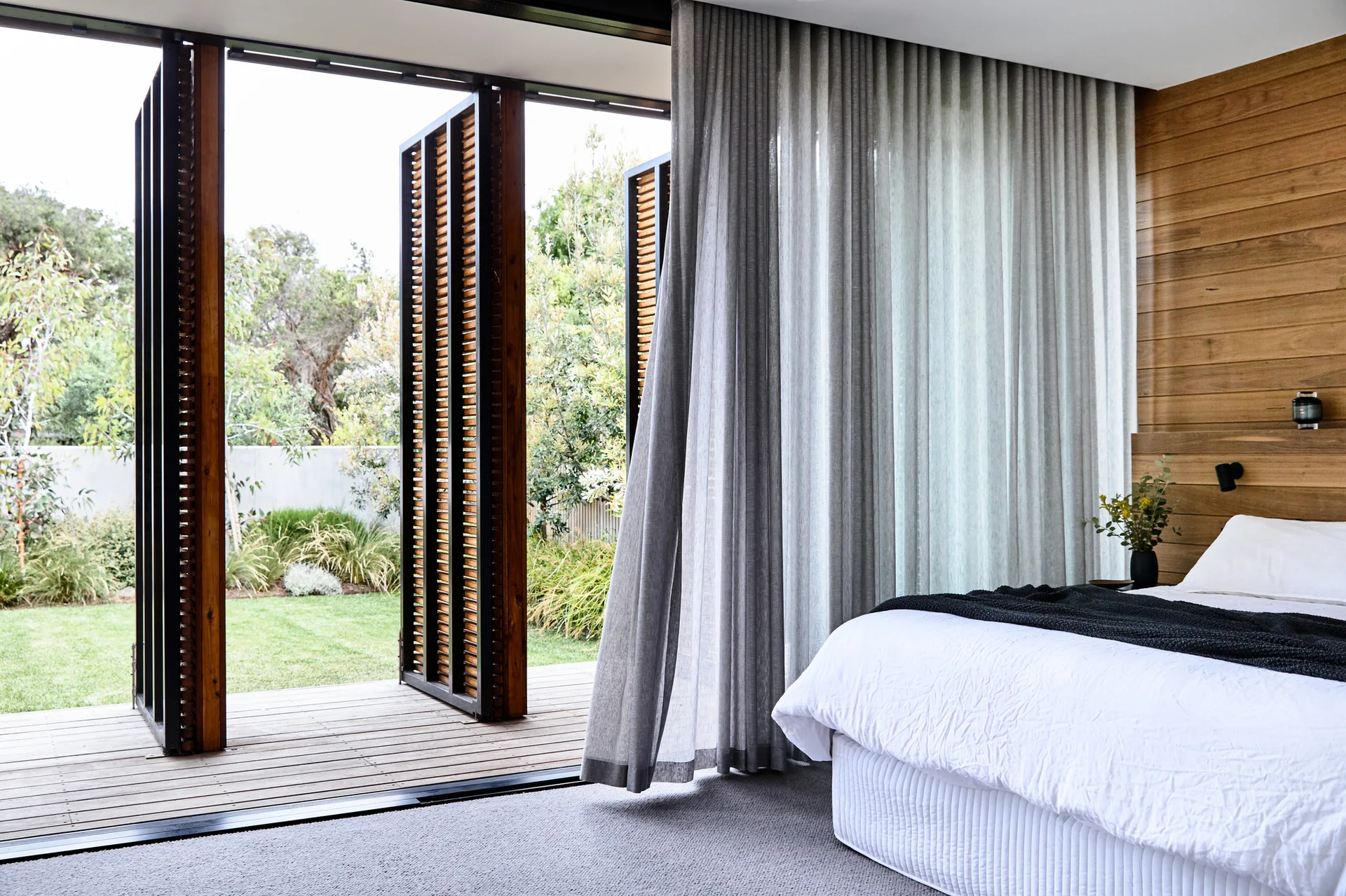 A modern bedroom with white bedding, a wood plank accent wall, and open louvered bi-fold doors leading to a wooden deck and green garden.