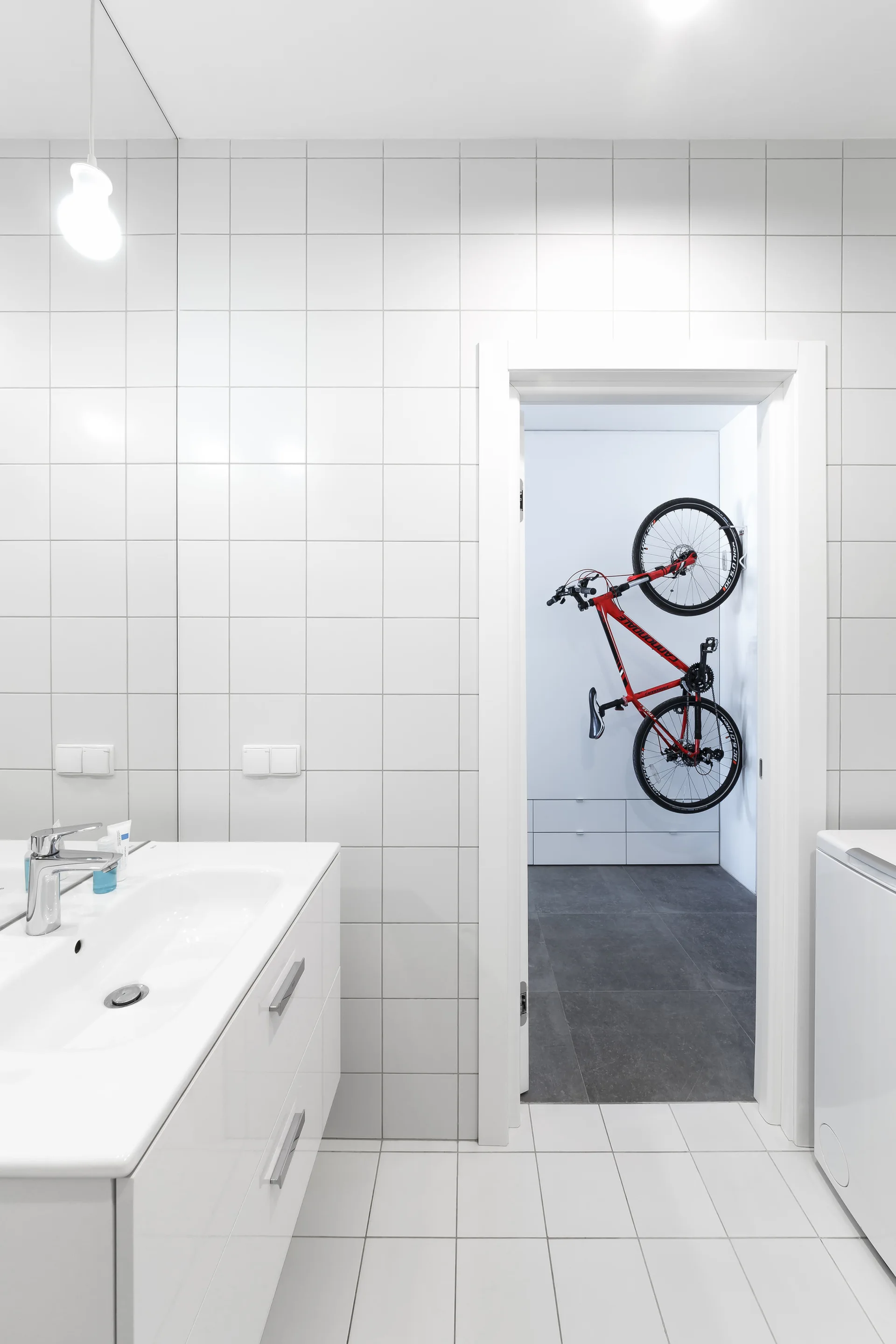 A white-tiled bathroom with a floating vanity looks through an open doorway into an adjacent room where a red bicycle is mounted on the wall.