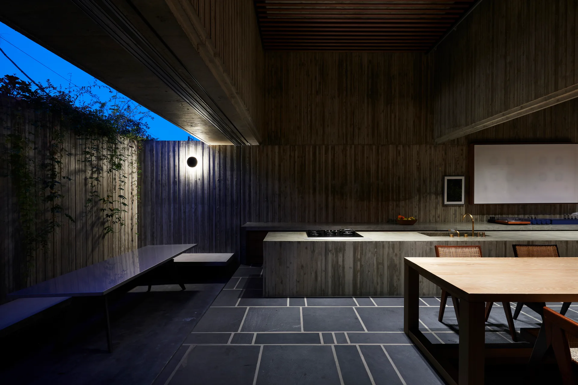 A modern minimalist kitchen and dining space with textured board-formed concrete walls, a concrete island, and a dark stone tiled floor.