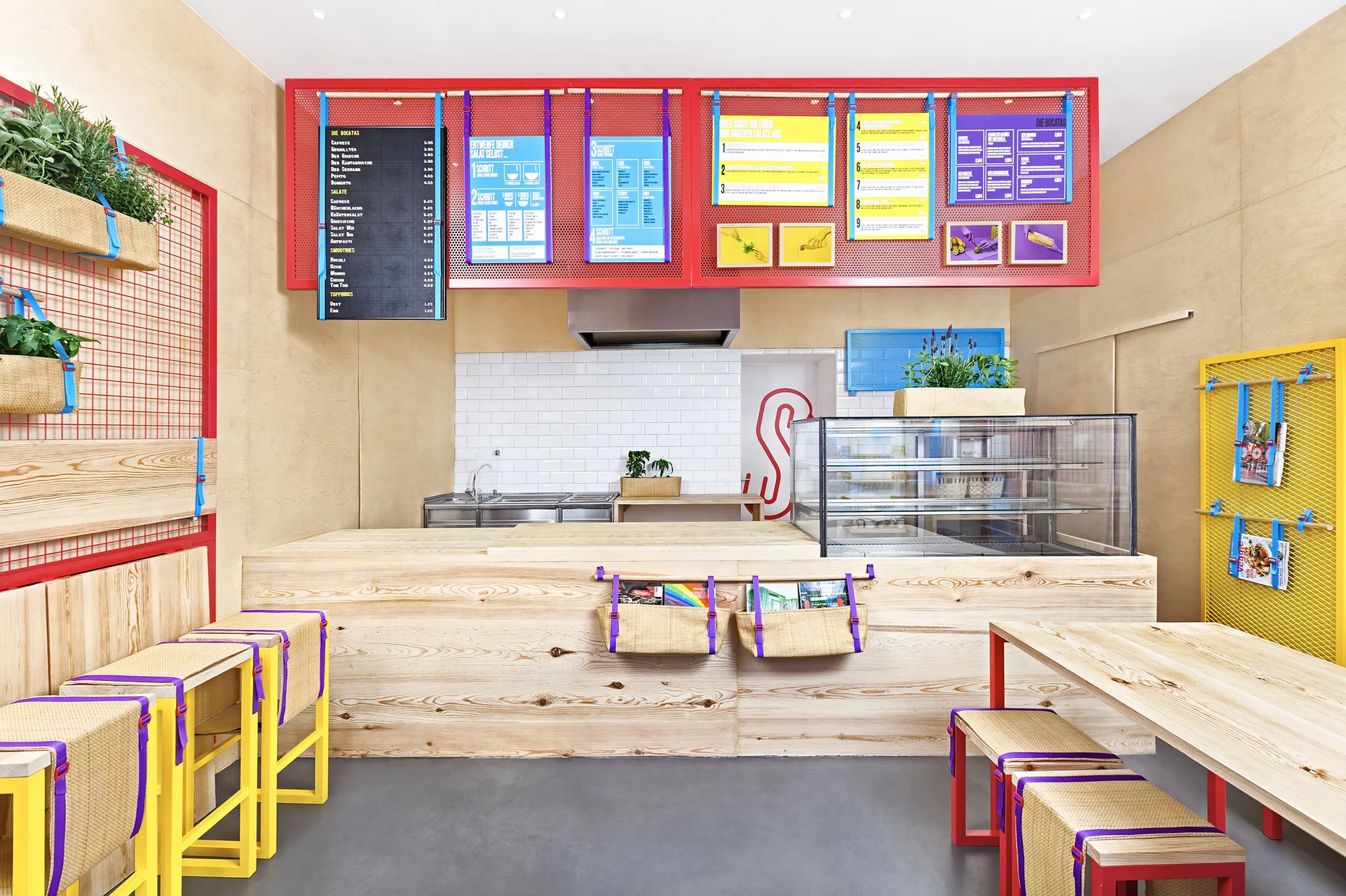 A modern commercial cafe featuring a light plywood service counter, vibrant red and yellow metal grids, and matching wooden seating.