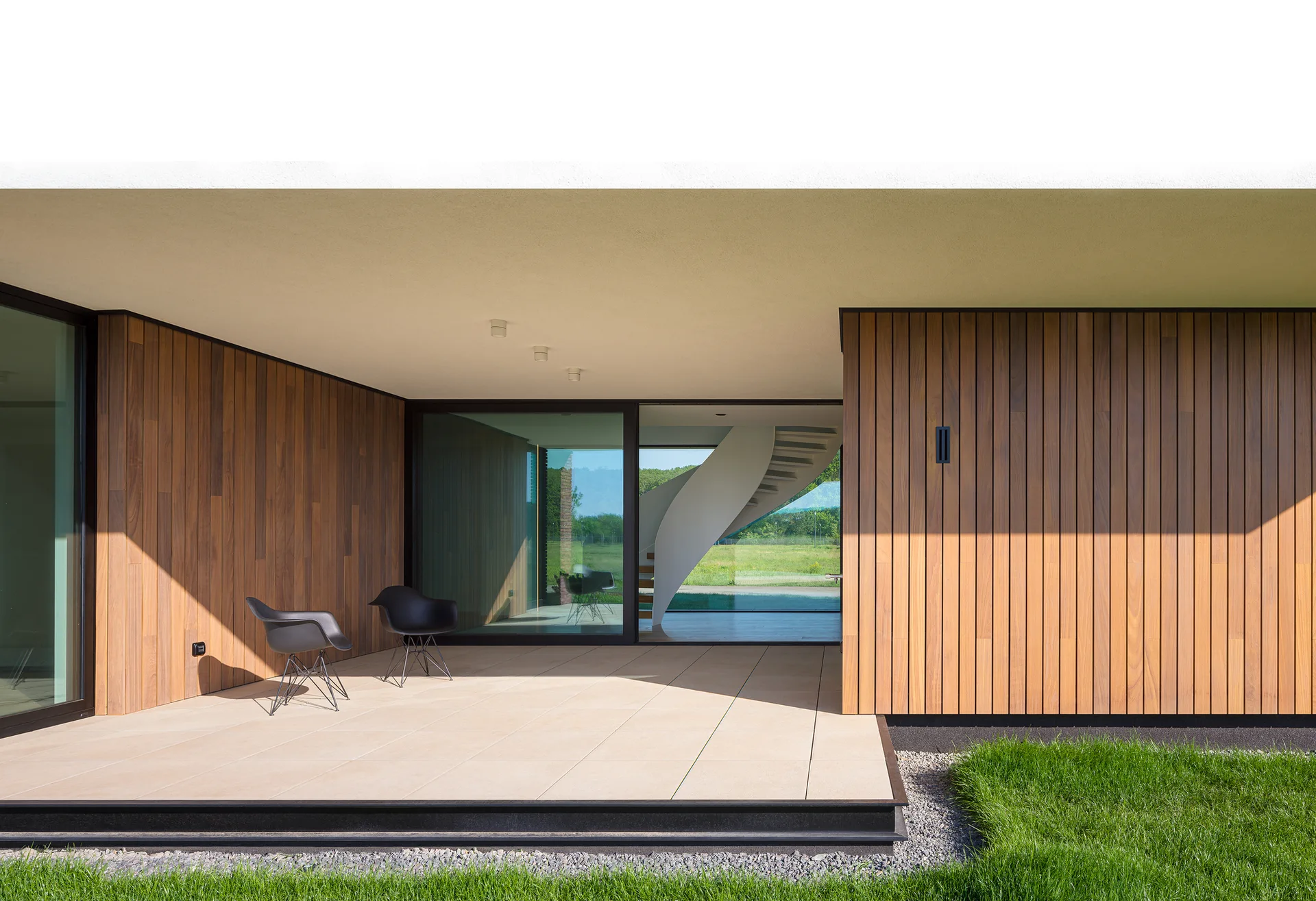 A covered patio with vertical wood siding and beige tiled floors features two black chairs and a sliding glass door revealing an interior curved staircase.