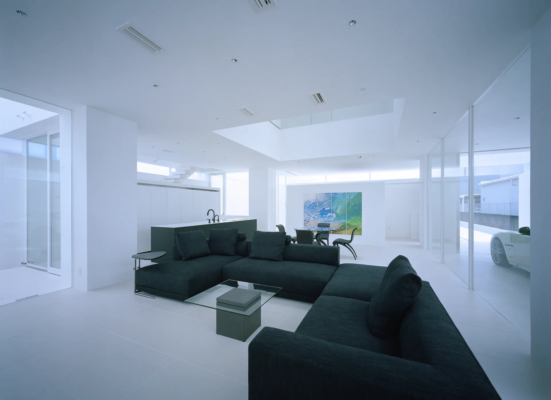 A minimalist open-concept white living room features a large charcoal sectional sofa, a glass coffee table, and floor-to-ceiling glass partitions.