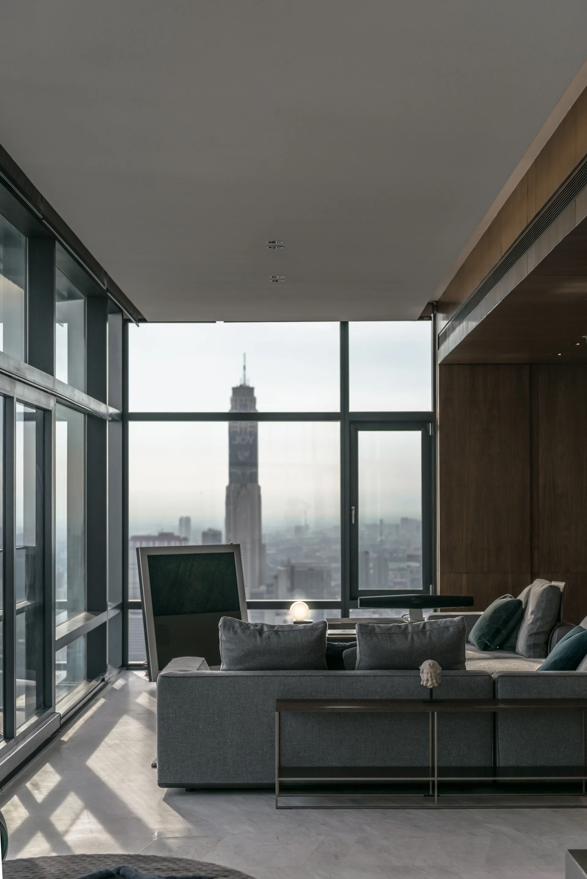 A modern living room features a grey sectional sofa, a slim console table, and a dark wood paneled wall next to large windows with a city view.