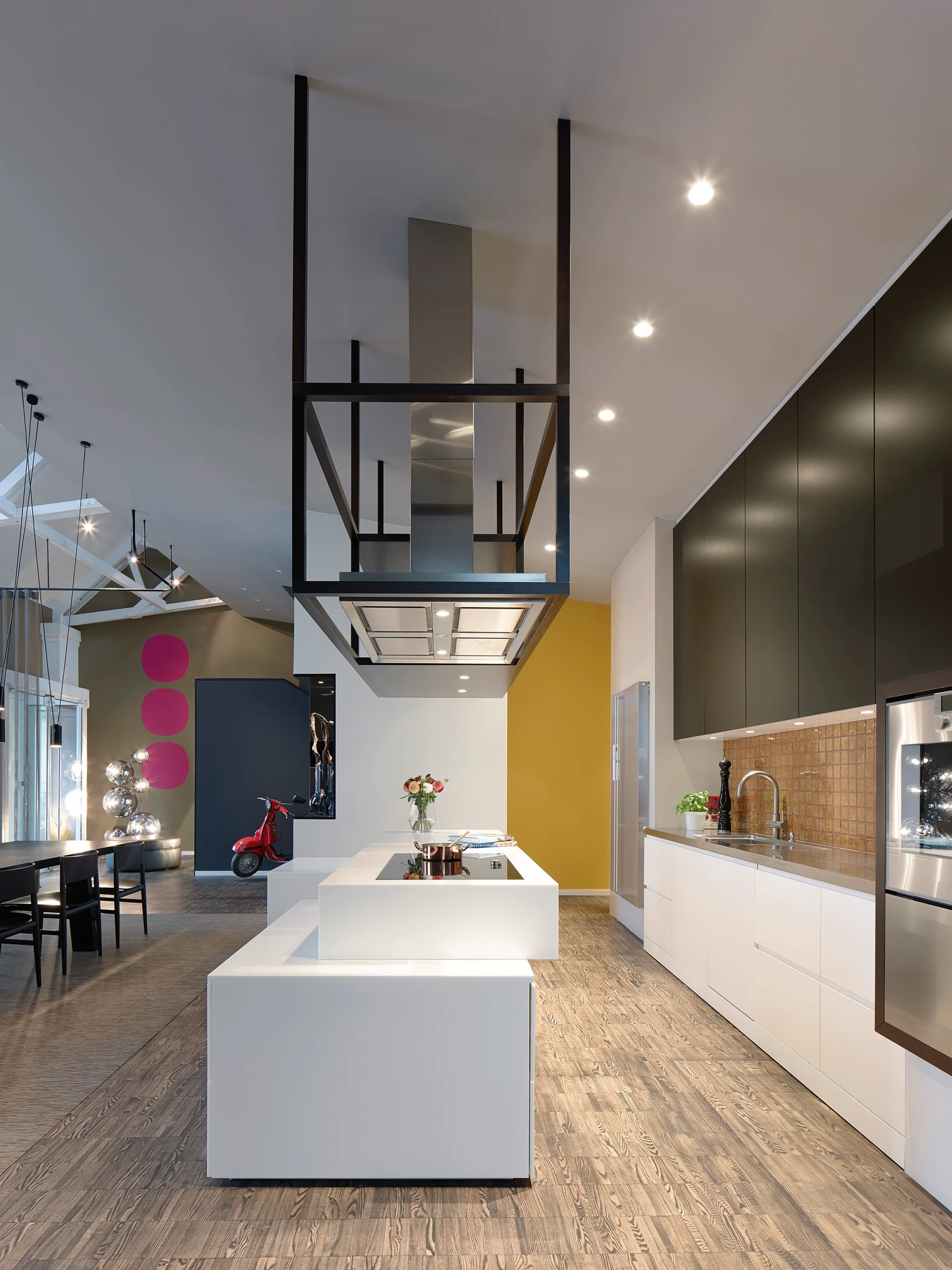 A modern open-concept kitchen features a tiered white island beneath a black sculptural range hood, flanked by minimalist black and white cabinets.