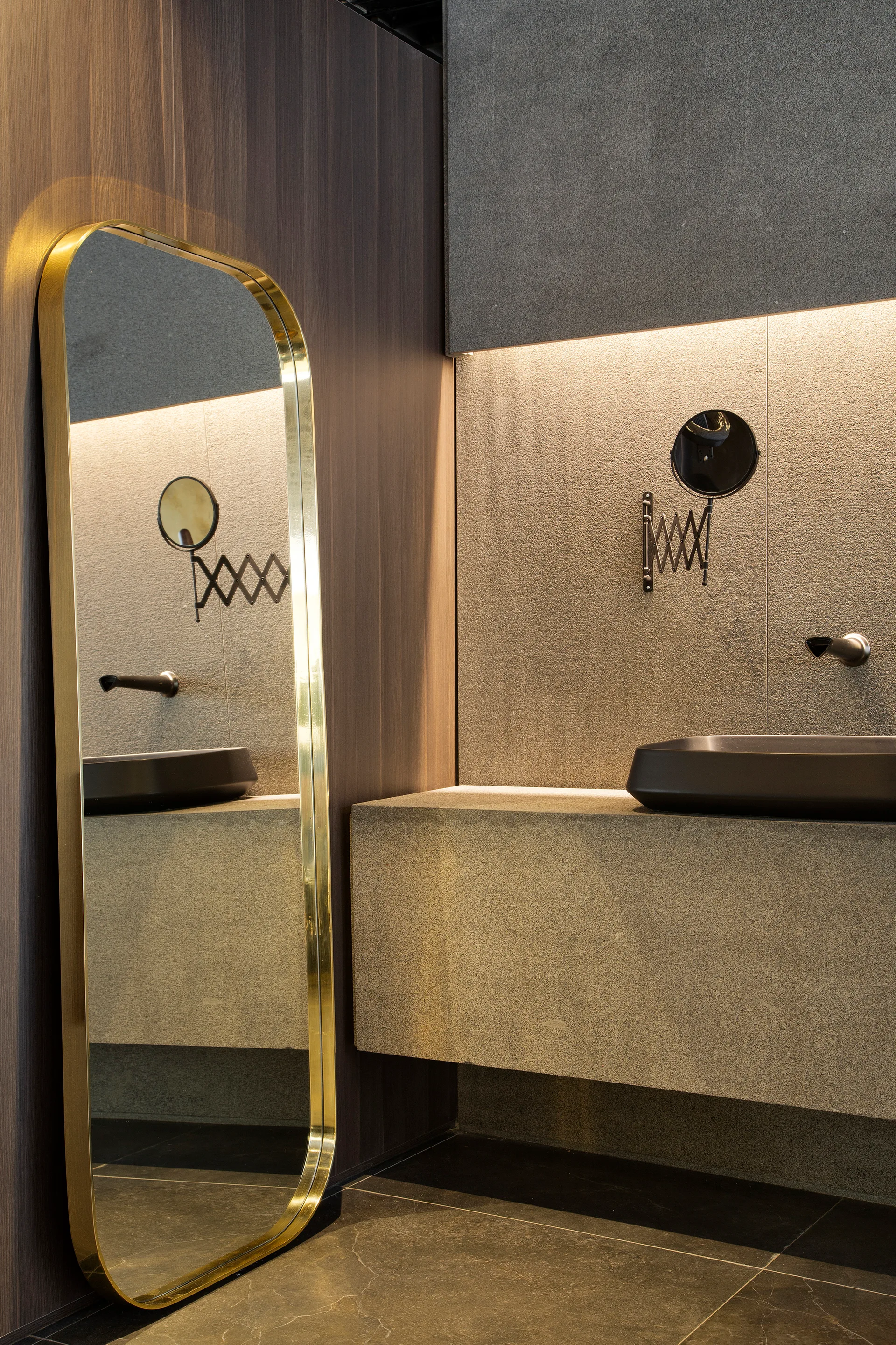A modern bathroom vanity area with a floating stone sink next to a tall gold-framed floor mirror against textured stone and wood walls.