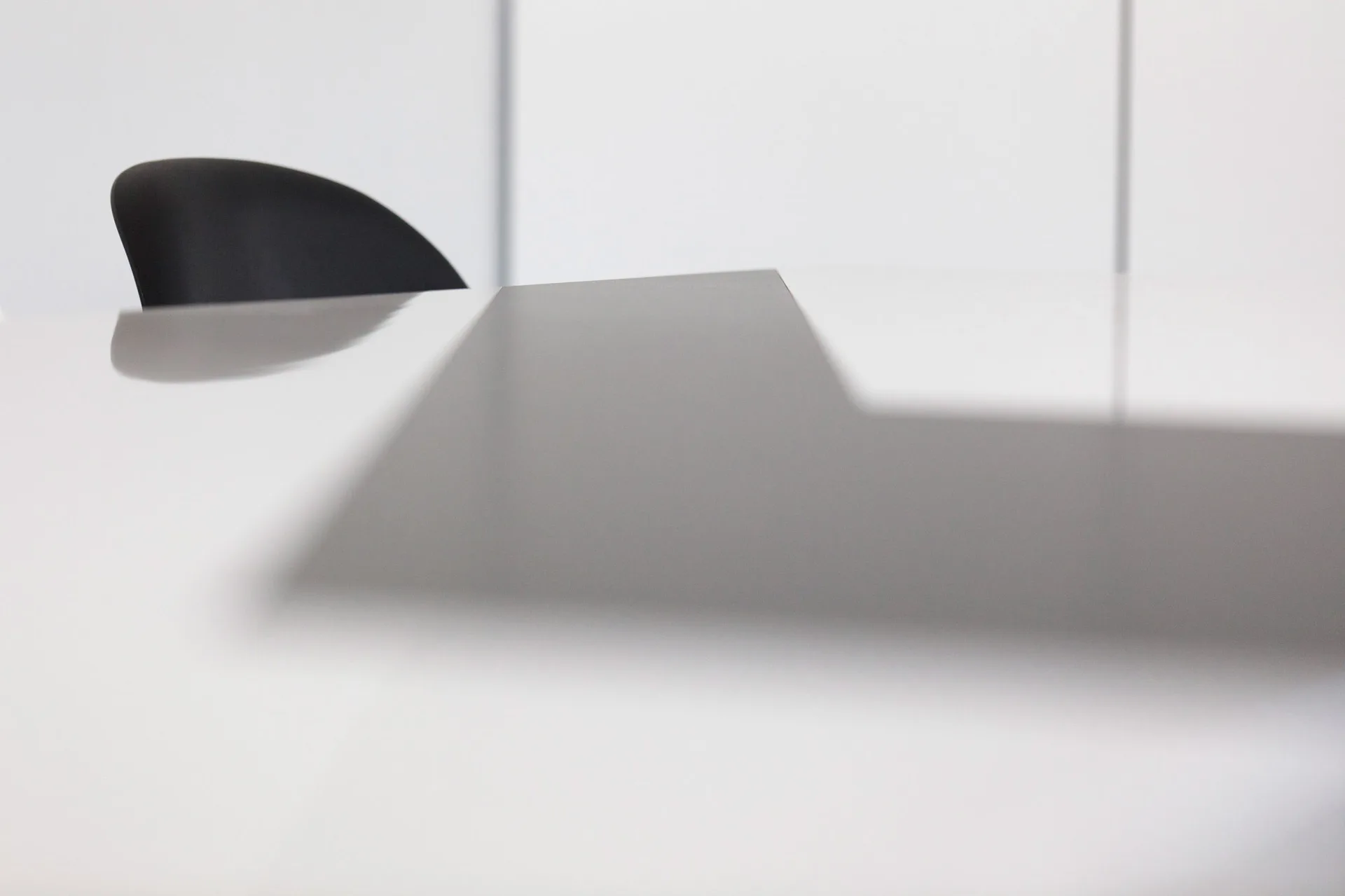 A close-up view of a smooth white table surface with a dark black chair back silhouette against a white wall.