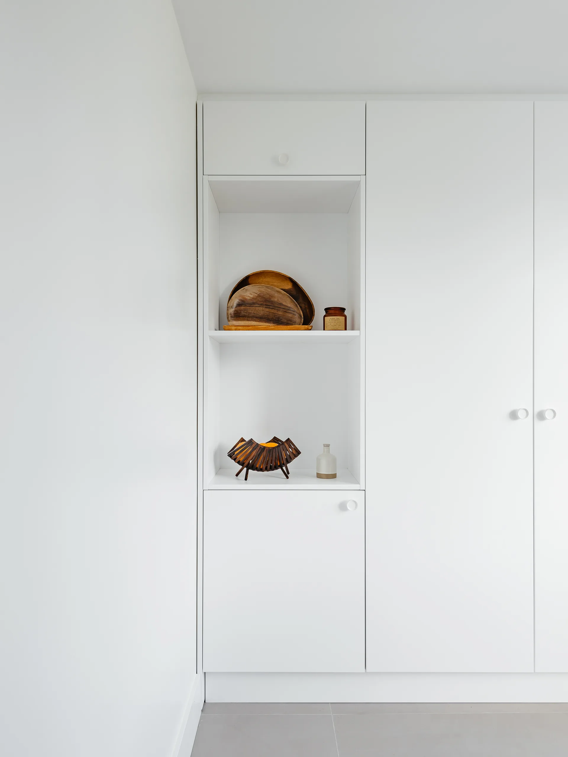 White minimalist built-in cabinets feature a recessed open shelving niche displaying wooden bowls, a slatted wooden basket, and small decorative bottles.