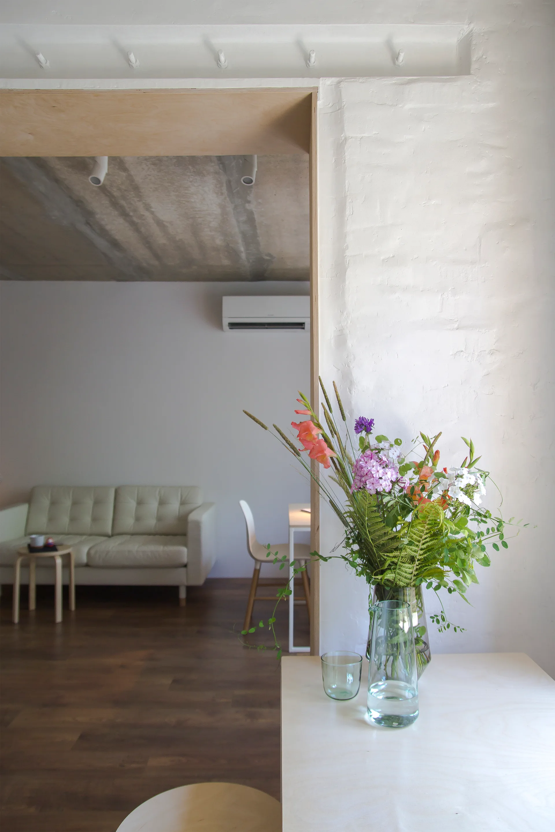 A view through a plywood-framed doorway of a bright living room featuring a white leather sofa, concrete ceiling, and a flower vase in the foreground.