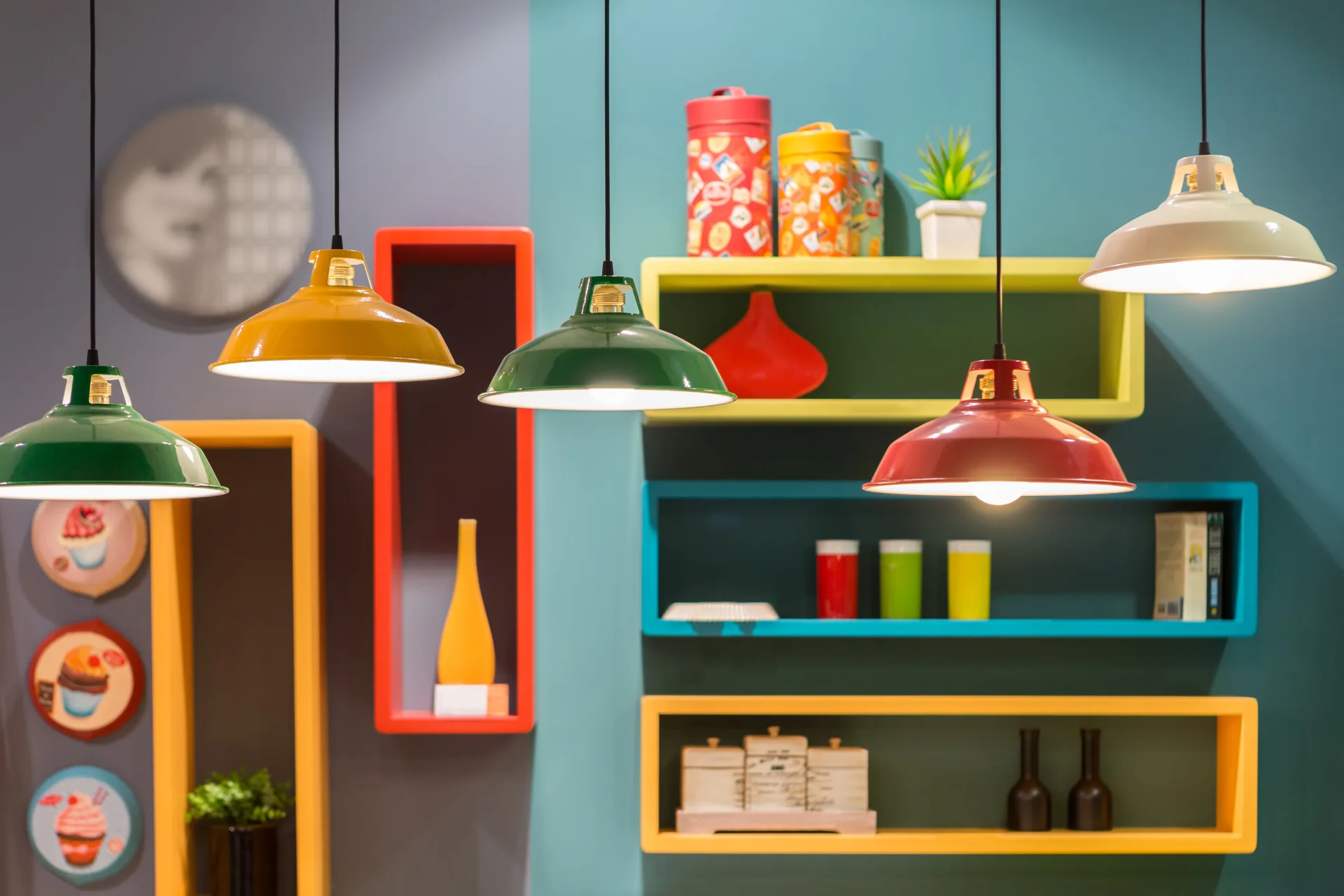 Several colorful dome pendant lights hang at varying heights in front of a wall featuring bright blue, yellow, and red box shelves.