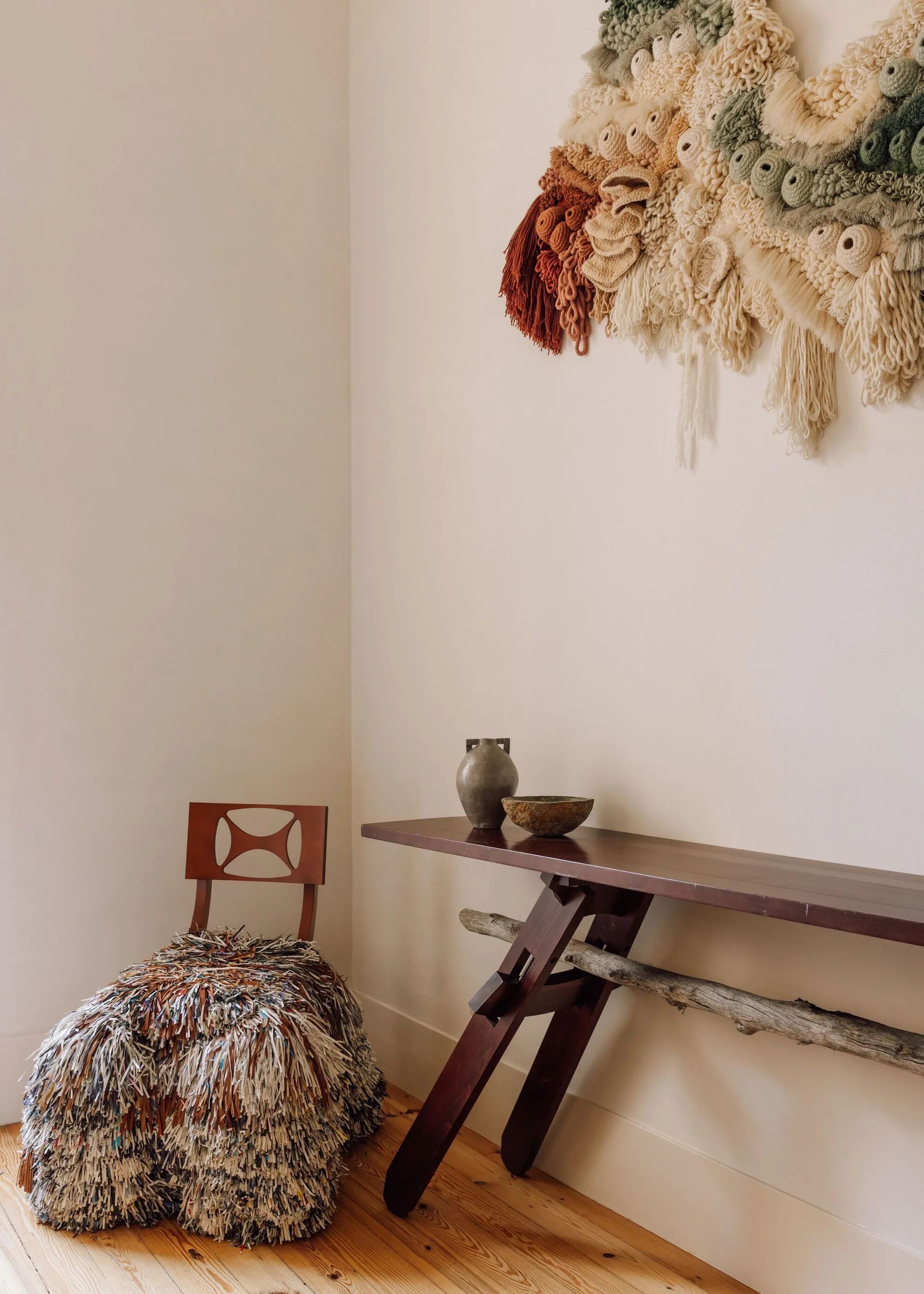 A dark wood console table and a wooden chair with a bulky shaggy seat sit in a room corner beneath a large textured fiber wall hanging.