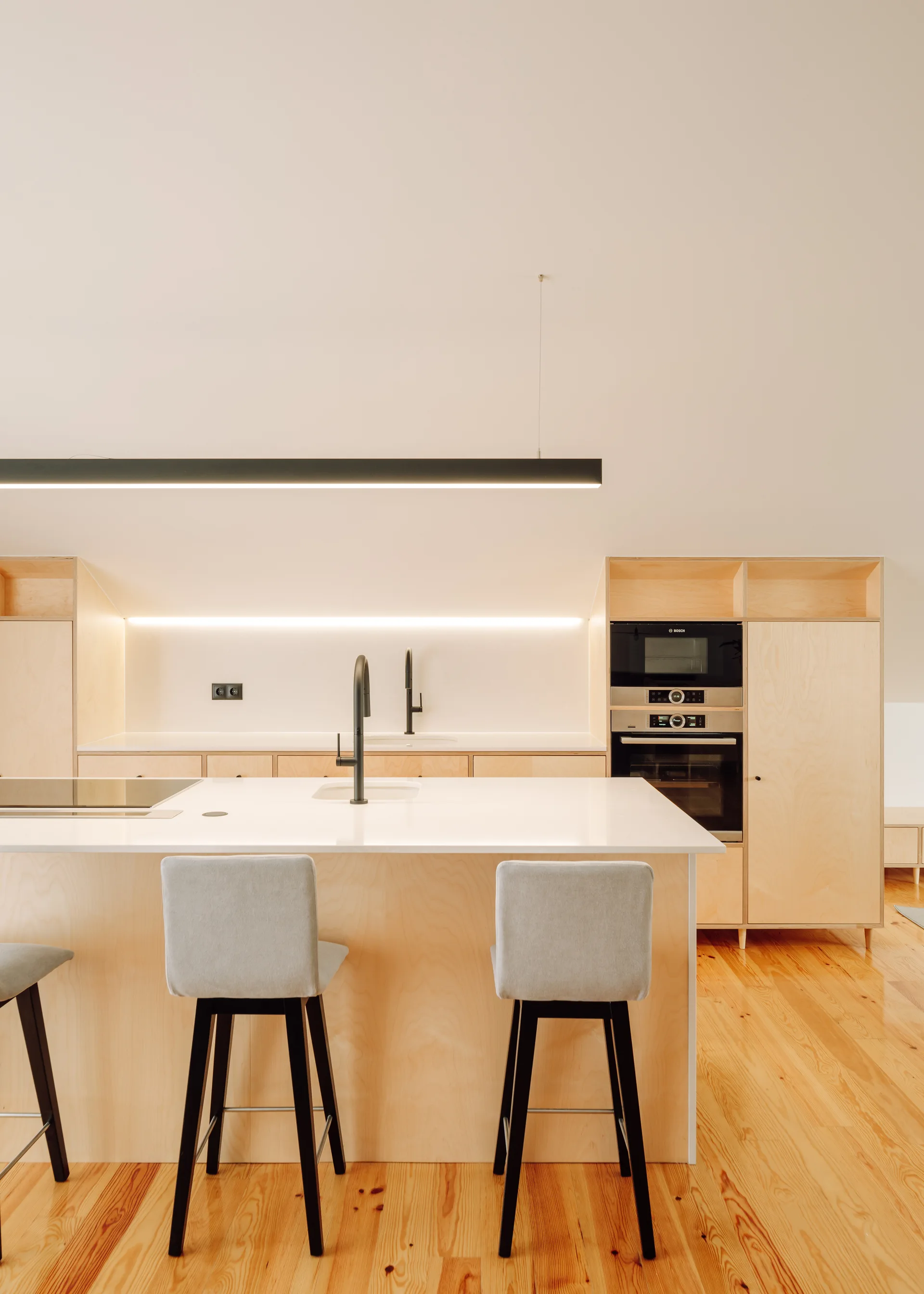 A minimalist Scandinavian kitchen featuring light plywood cabinetry, a white countertop island with gray stools, and a black linear pendant light.