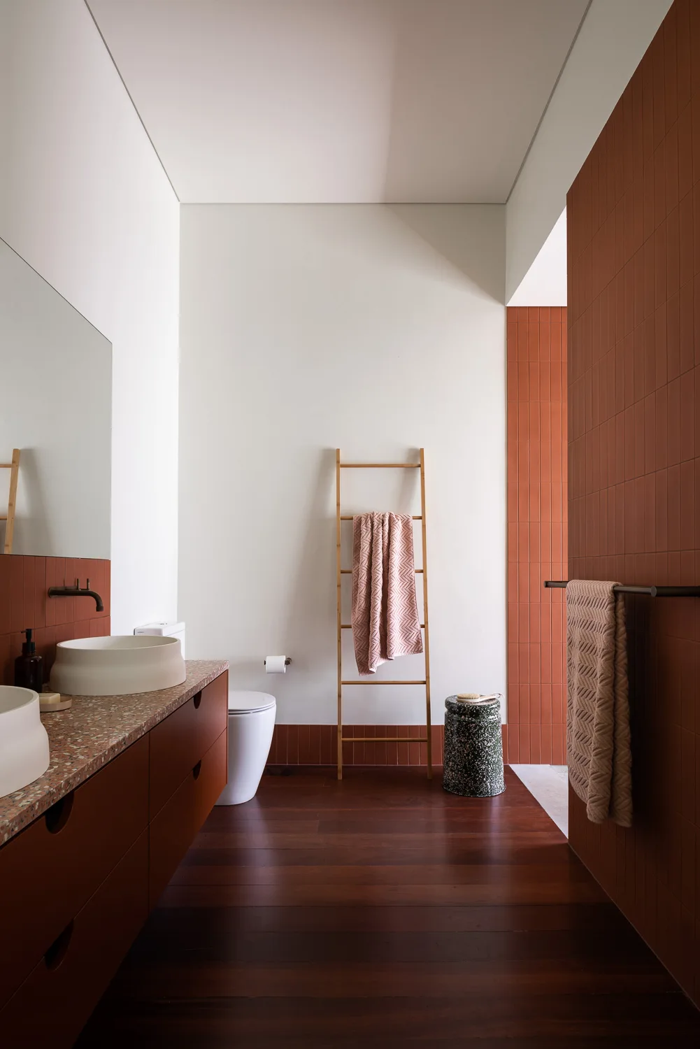 A minimalist bathroom with terracotta wall tiles, a dark wood vanity, white vessel sinks, and a wooden towel ladder against a white wall.