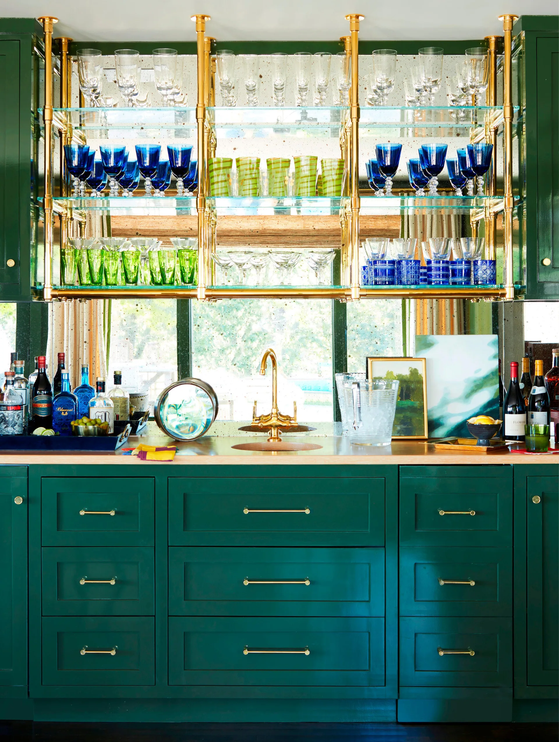 A home bar features forest green base cabinetry beneath suspended brass and glass shelving stacked with colorful glassware against an antique mirror backdrop.