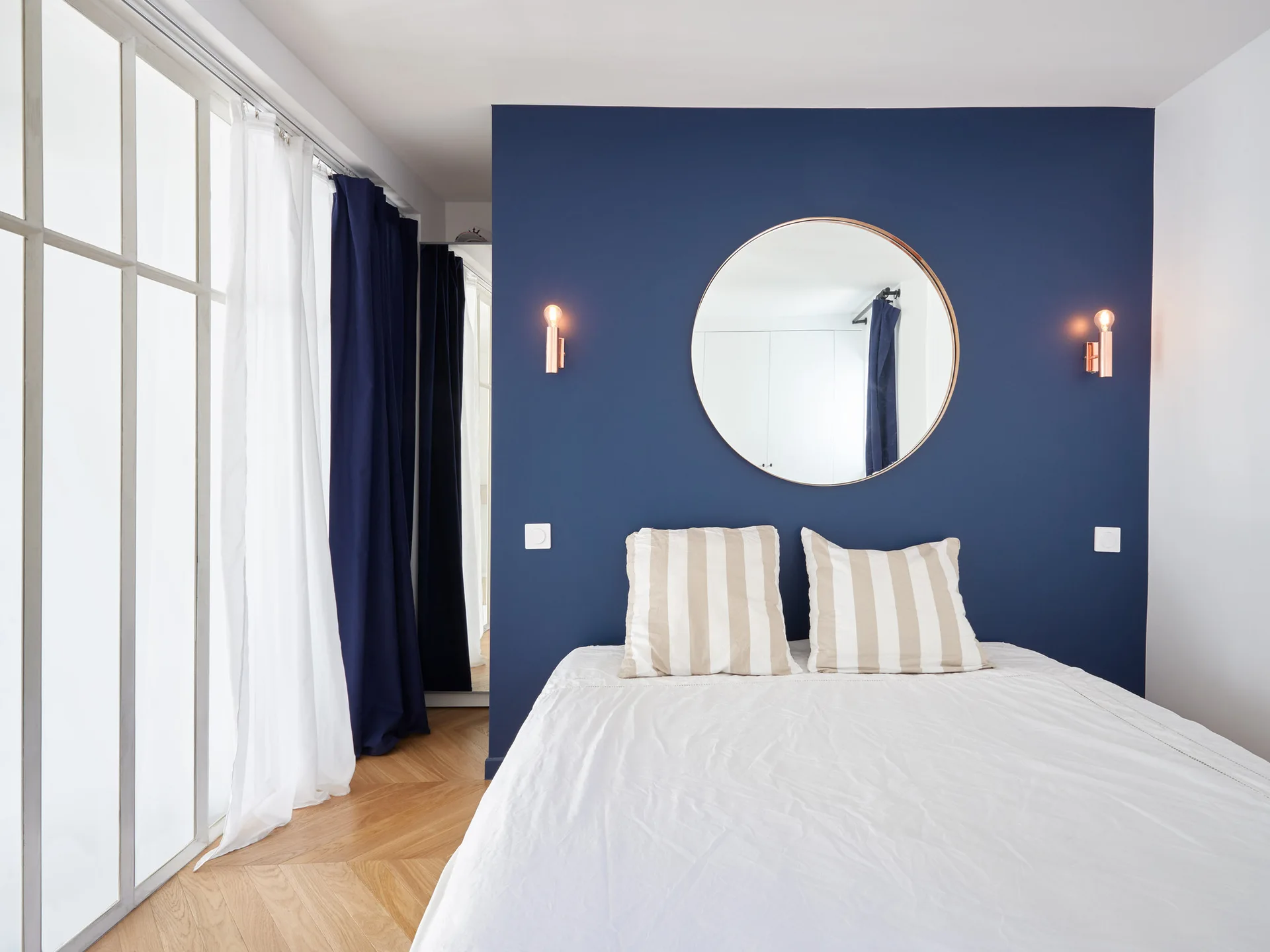 A modern bedroom featuring a bed with striped pillows against a dark navy blue accent wall with a round mirror and symmetrical wall sconces.