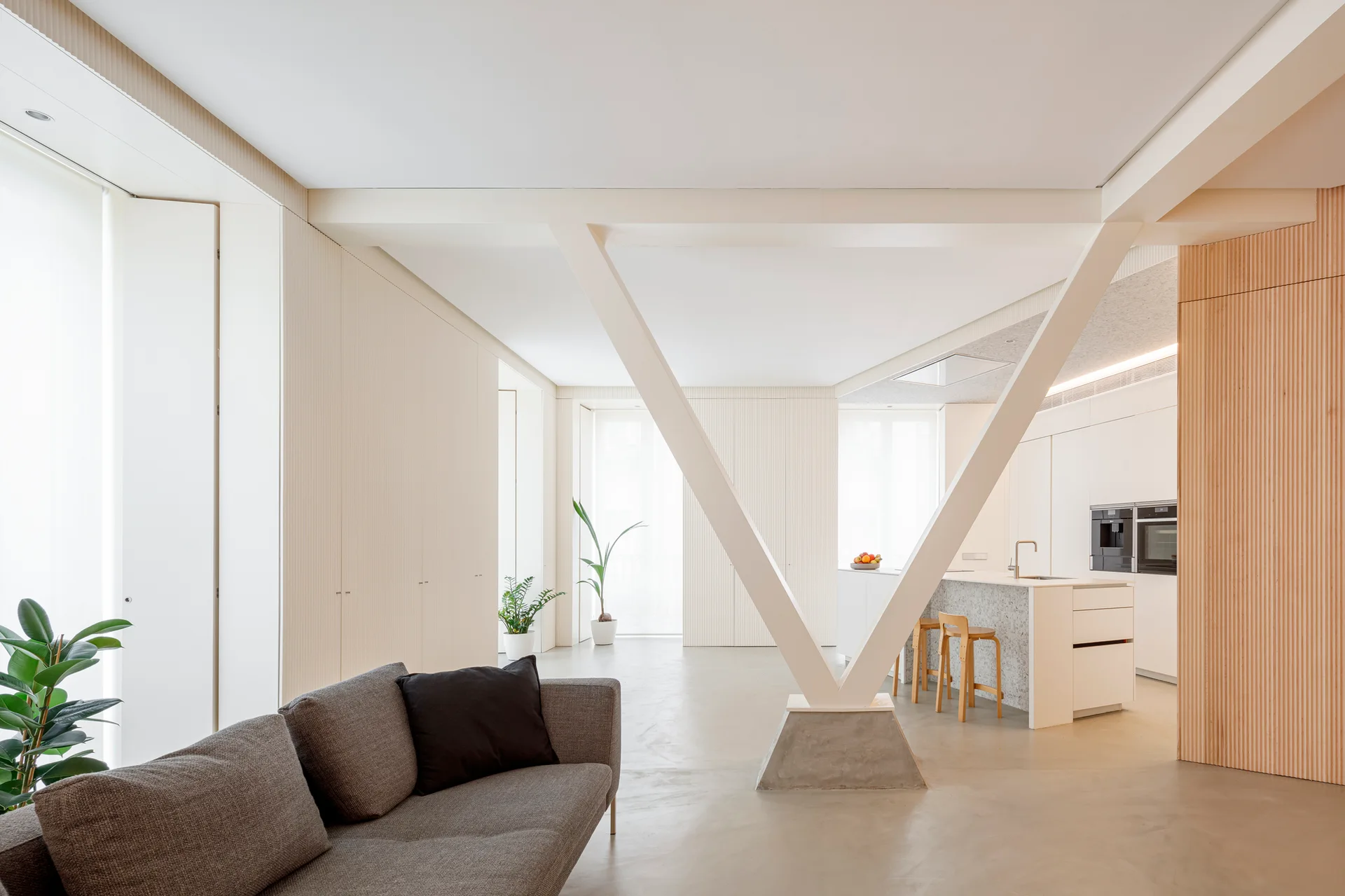 A modern open-plan living area and kitchen featuring a grey sofa, a prominent V-shaped structural beam, and fluted wood wall panels.