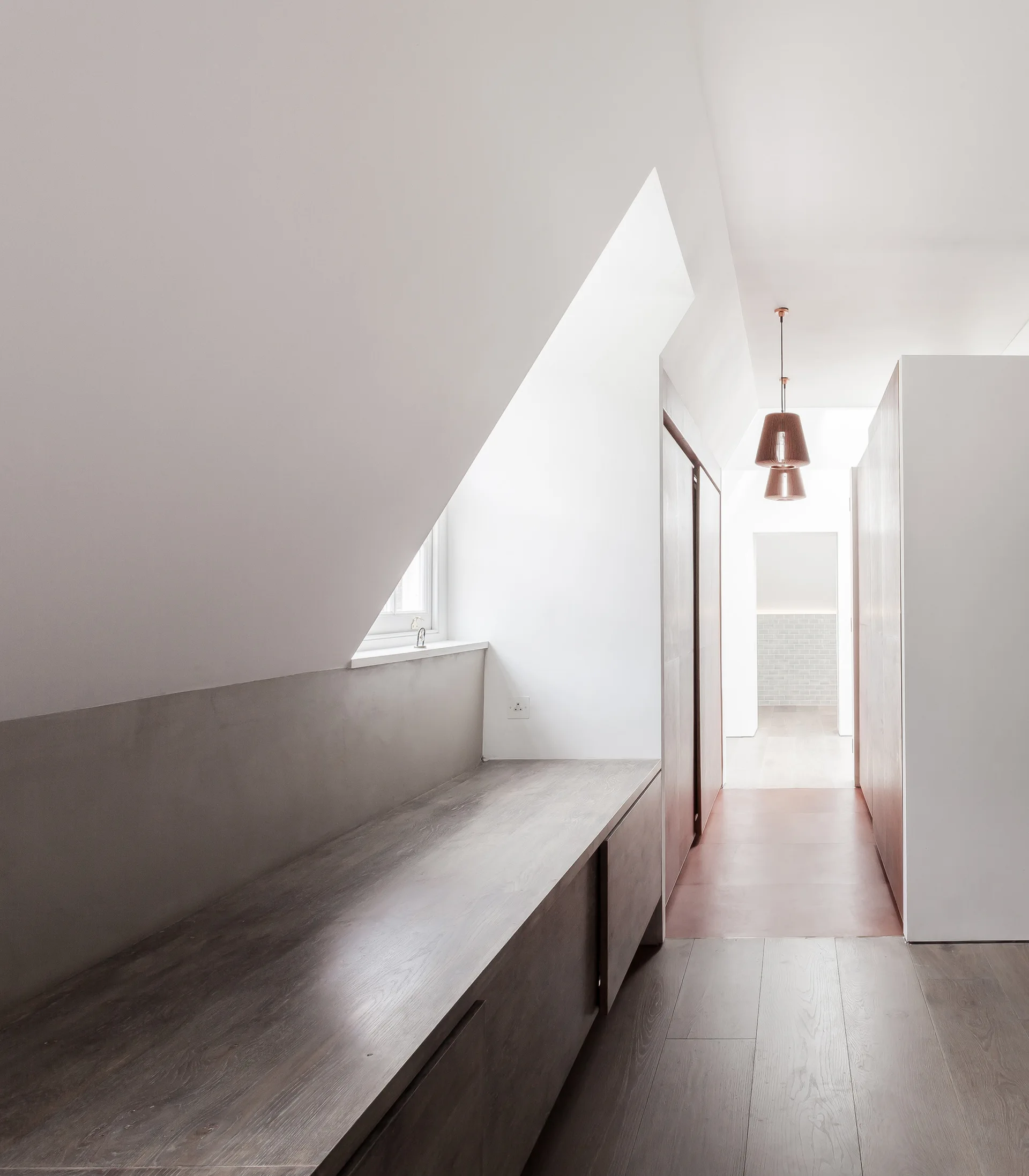 A minimalist attic hallway features a low dark wood built-in storage bench beneath a sloped ceiling and warm copper pendant lights.