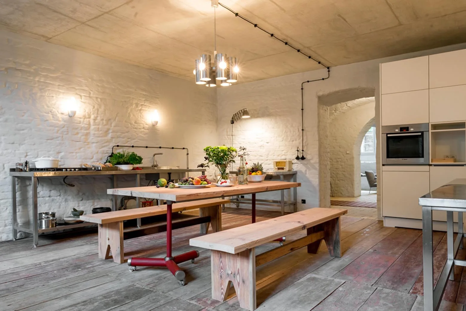 An industrial dining space features a wooden table with benches, white painted brick walls, a concrete ceiling, and a stainless steel kitchen counter.