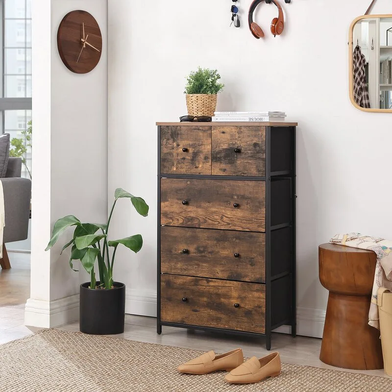 A five-drawer wood and metal chest of drawers stands against a white wall next to a potted houseplant and a cylindrical wooden stool on a woven rug.