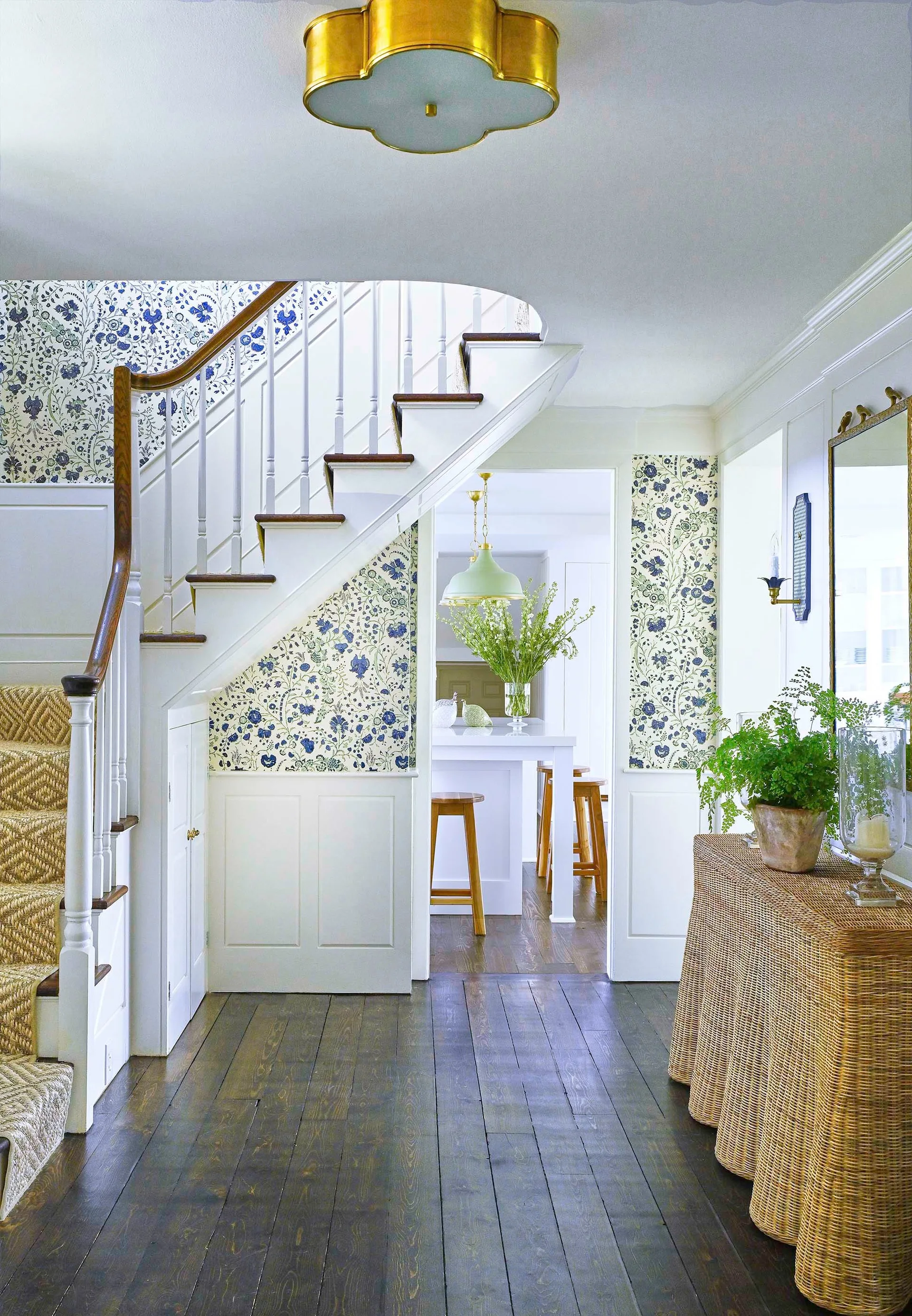 A bright foyer features blue and white floral wallpaper above white wainscoting, a wooden staircase, and a woven wicker console table on dark wood floors.