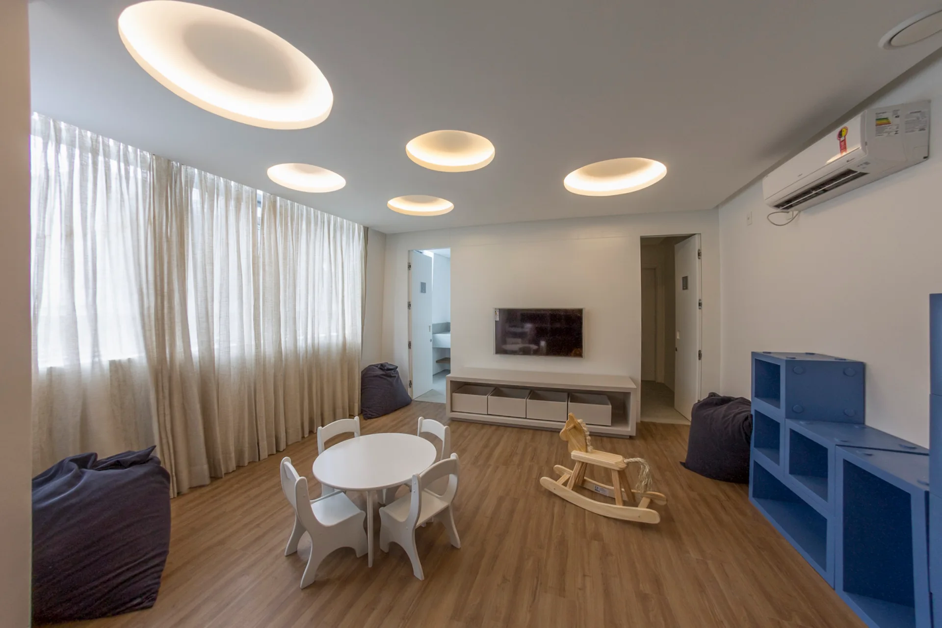 A bright children's playroom features white walls, circular ceiling lights, a small round table with matching chairs, and wood flooring.