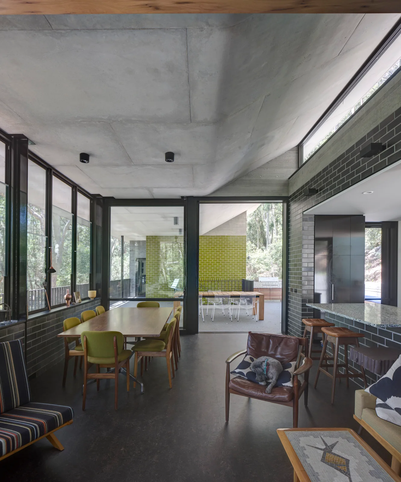 A modern open-concept dining room with an exposed concrete ceiling, a wood table surrounded by green chairs, and glass doors opening to a patio.