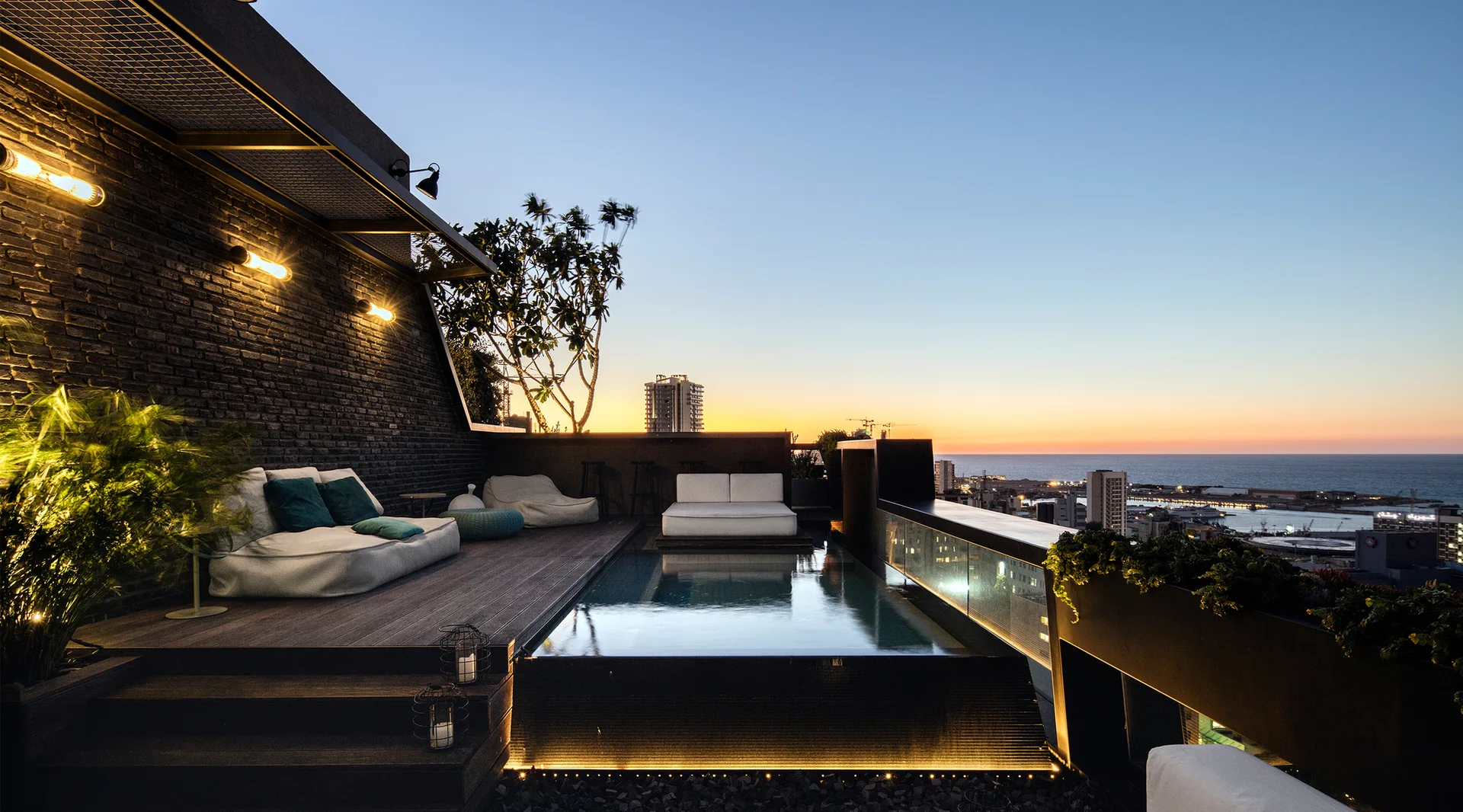 A modern rooftop terrace with a reflective infinity pool, dark wood decking, low-profile white sofas, and a dark brick wall during sunset.