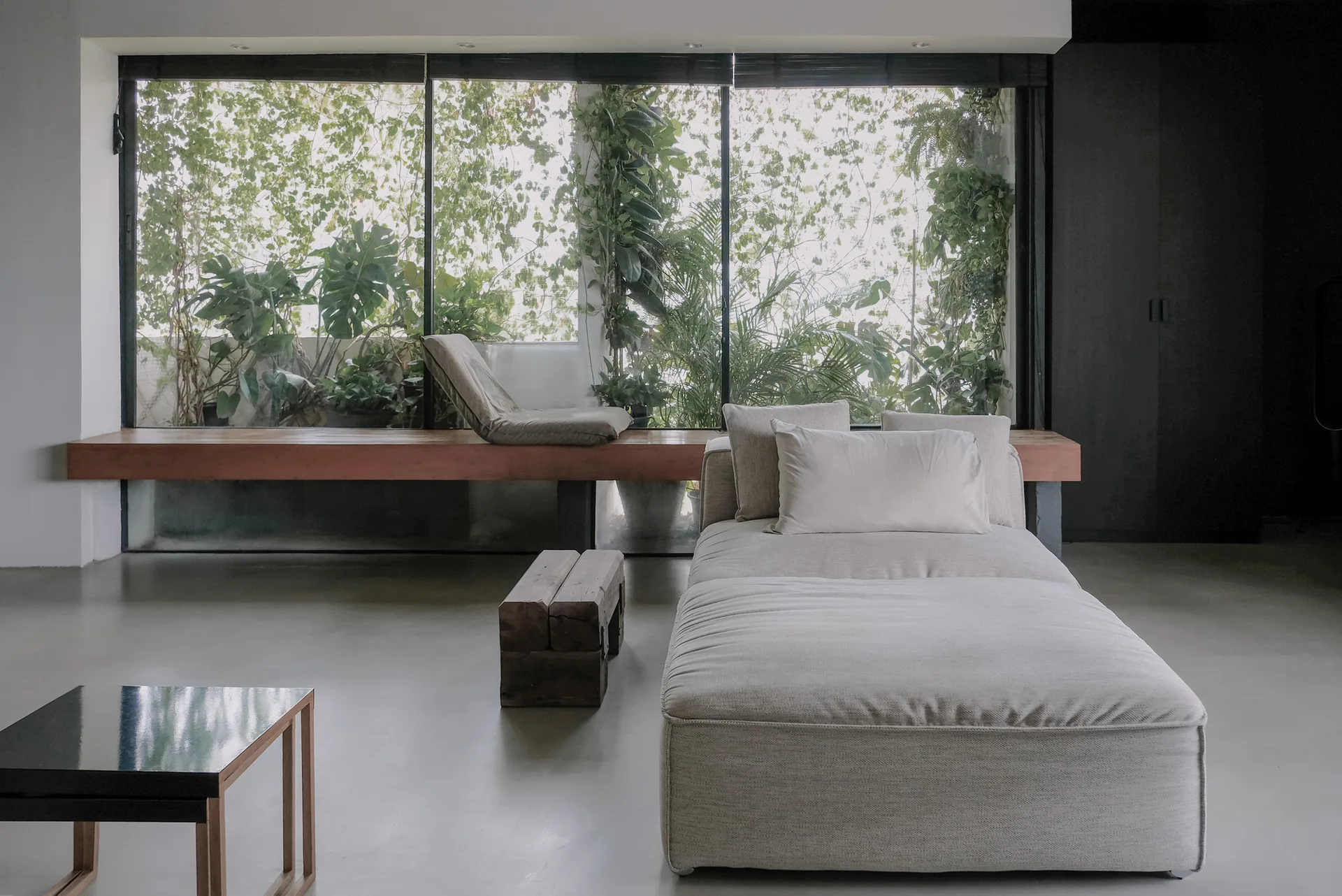 A minimalist living room featuring a beige chaise lounge, a concrete floor, and a long wooden bench in front of a large window filled with green plants.
