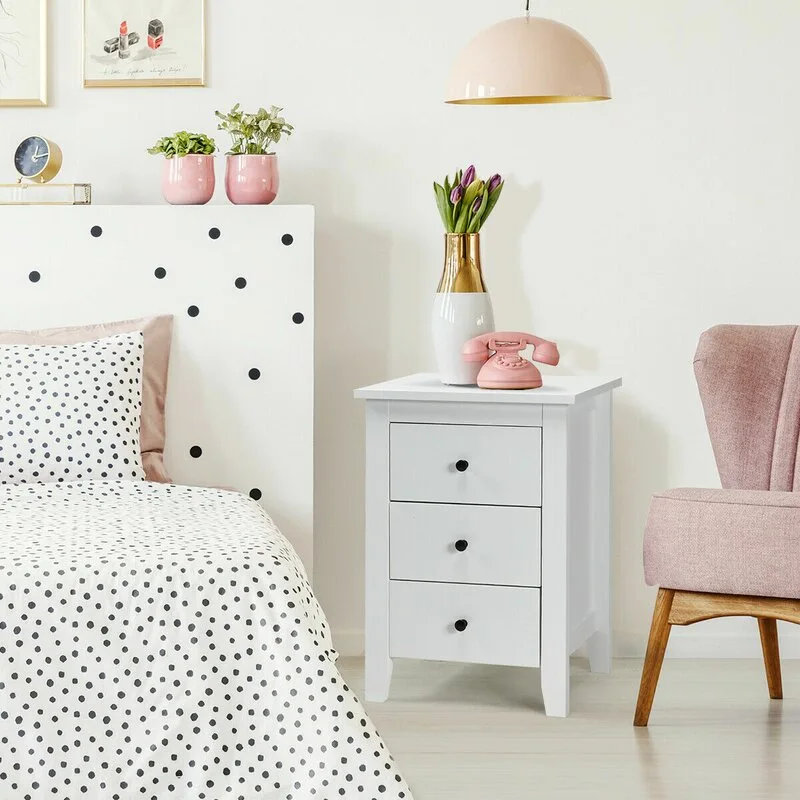 A white three-drawer nightstand stands next to a polka-dot bed and a soft pink armchair against a white wall.