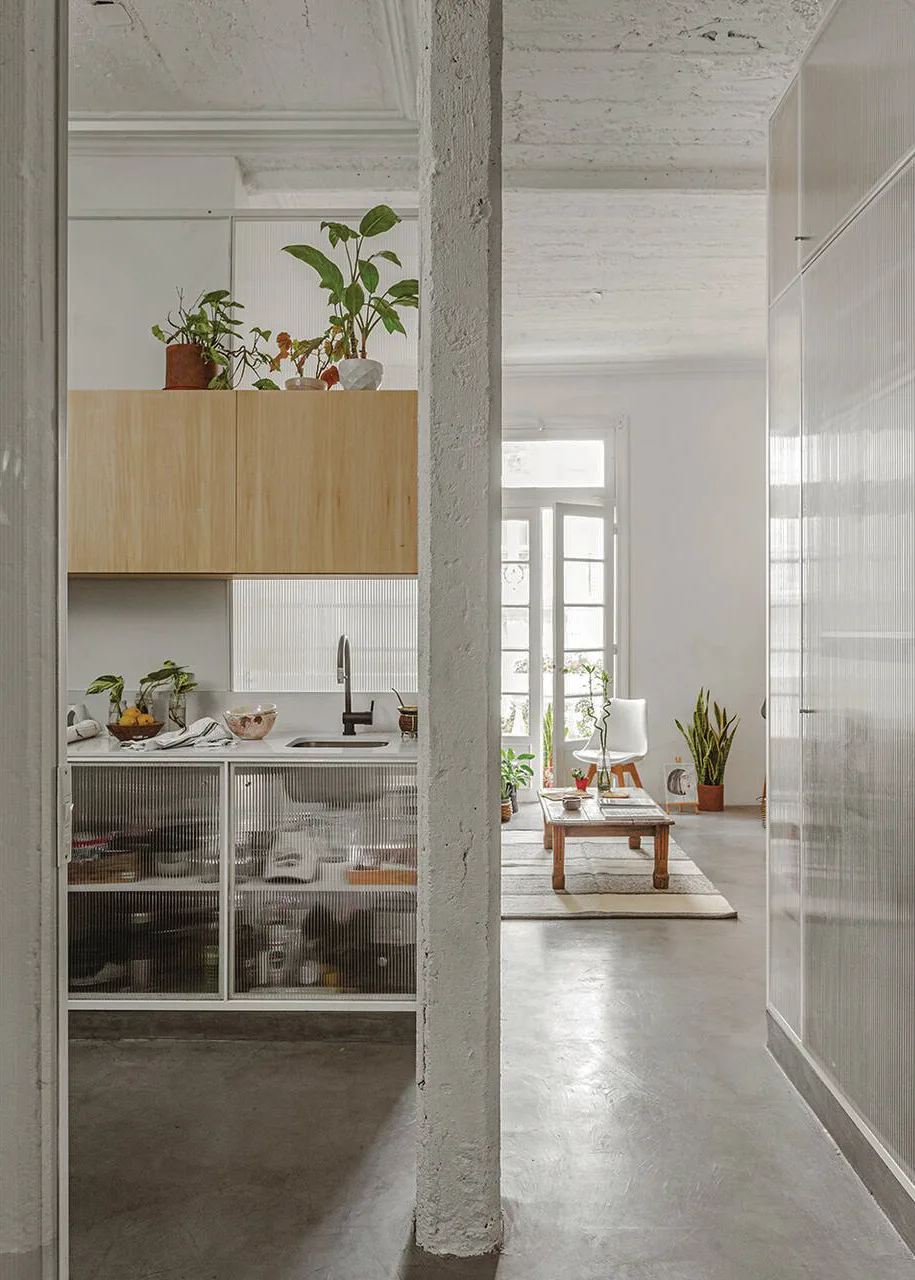 A modern open-plan kitchen and living space features fluted glass cabinets, light wood cupboards, a concrete pillar, and polished concrete floors.